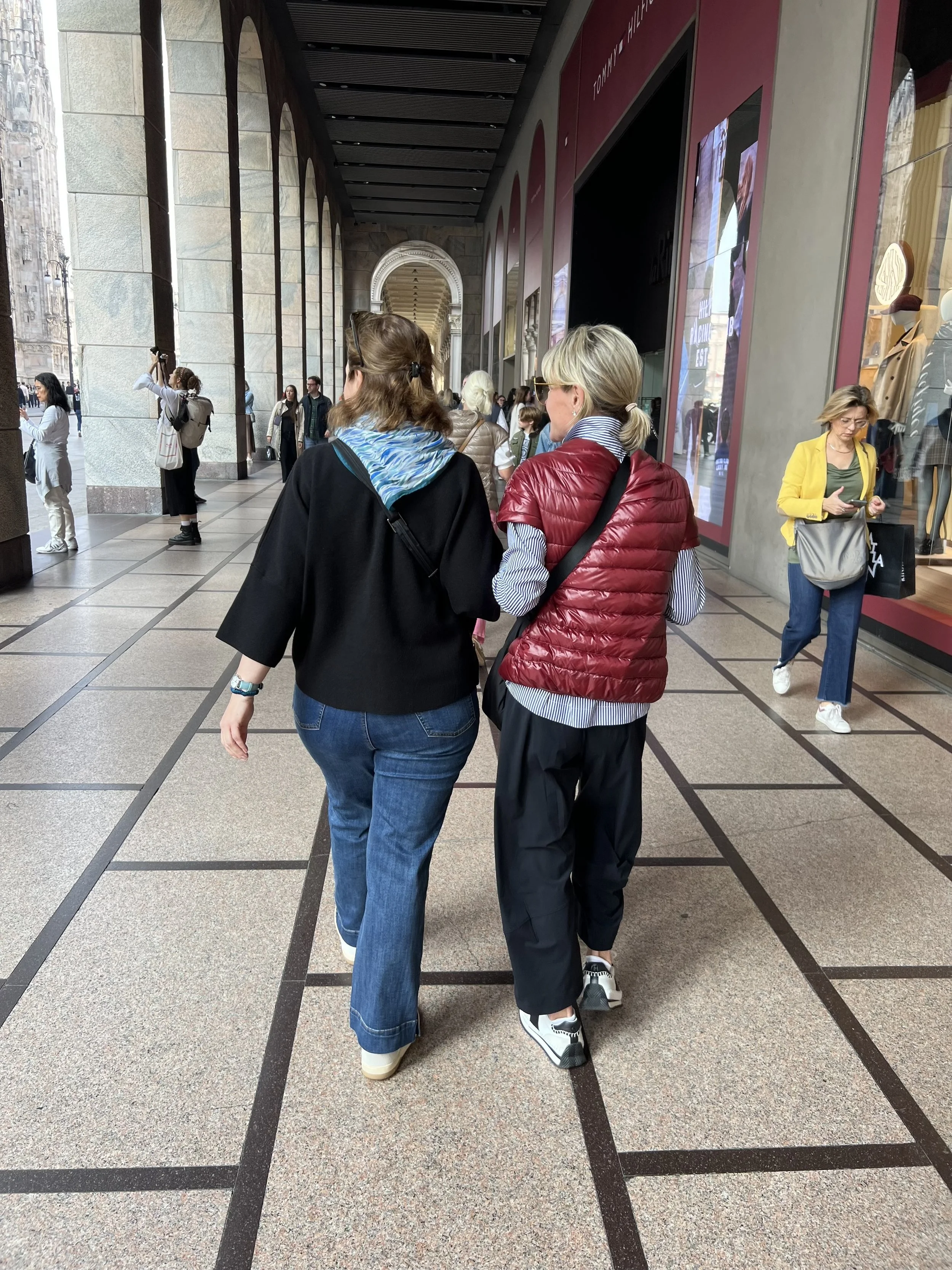 Two women walking side by side within a shopping mall or arcade, with people and shop windows around them.