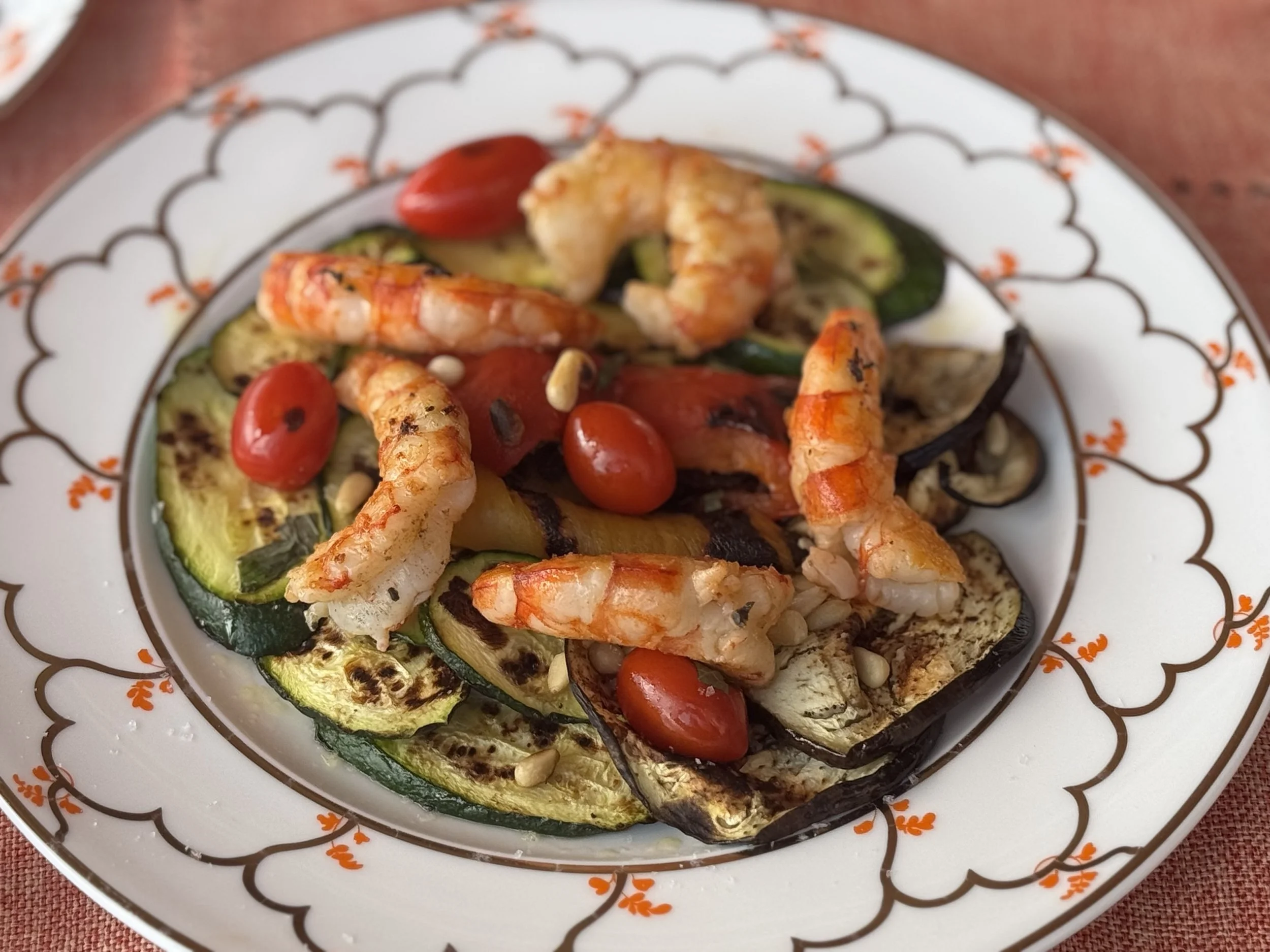 A plate of grilled zucchini, cherry tomatoes, grilled eggplant, and cooked shrimp served on a decorative white plate.