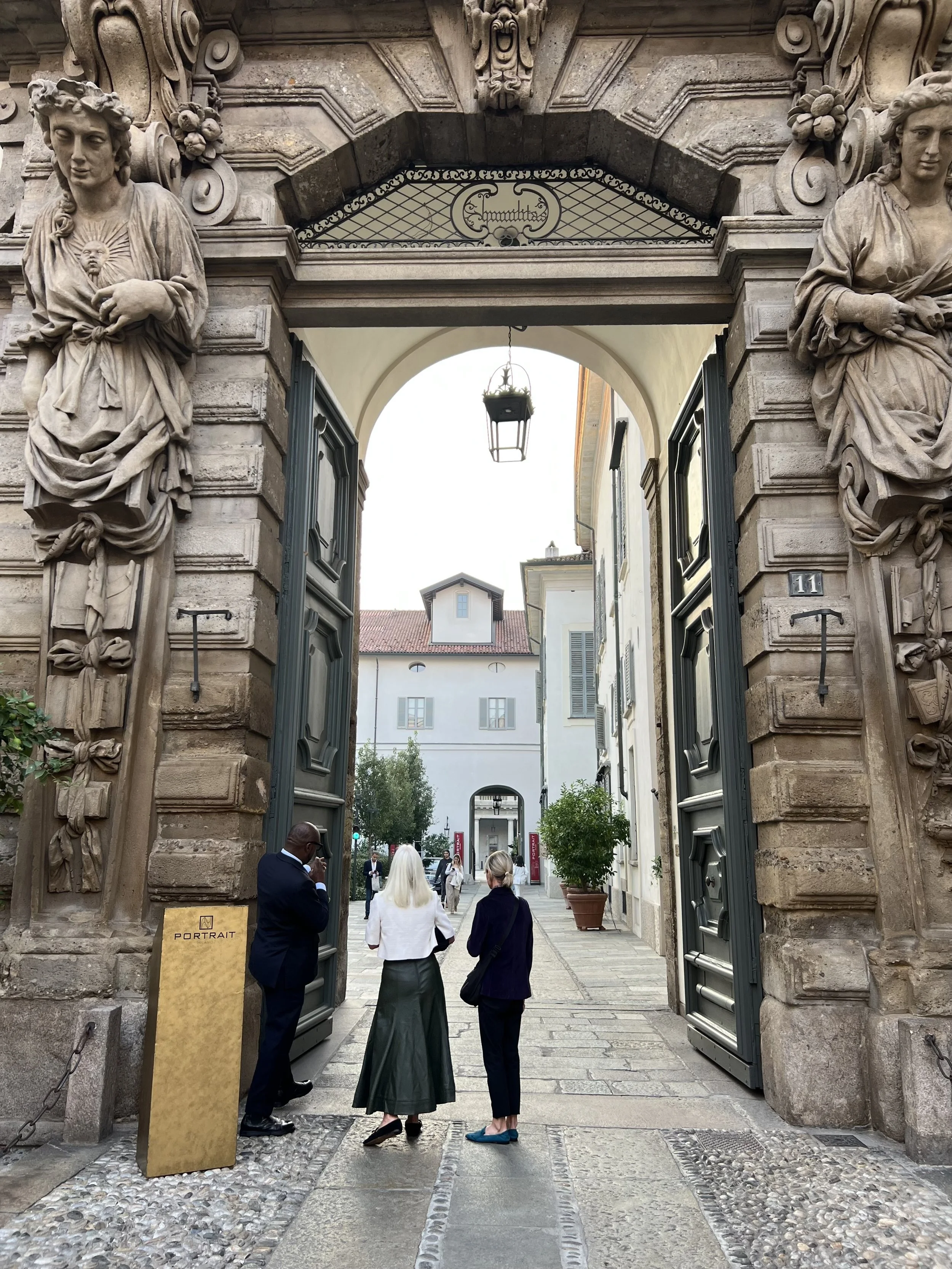People standing under an ornate stone archway entrance on a cobblestone street, with buildings and trees in the background.