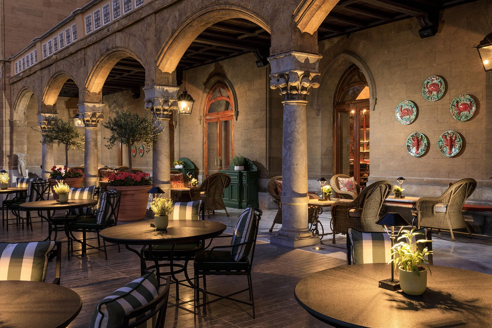 An outdoor patio area of a restaurant or cafe with tables and chairs, decorated with potted plants and surrounded by large stone arches and columns, illuminated by warm lighting at dusk.