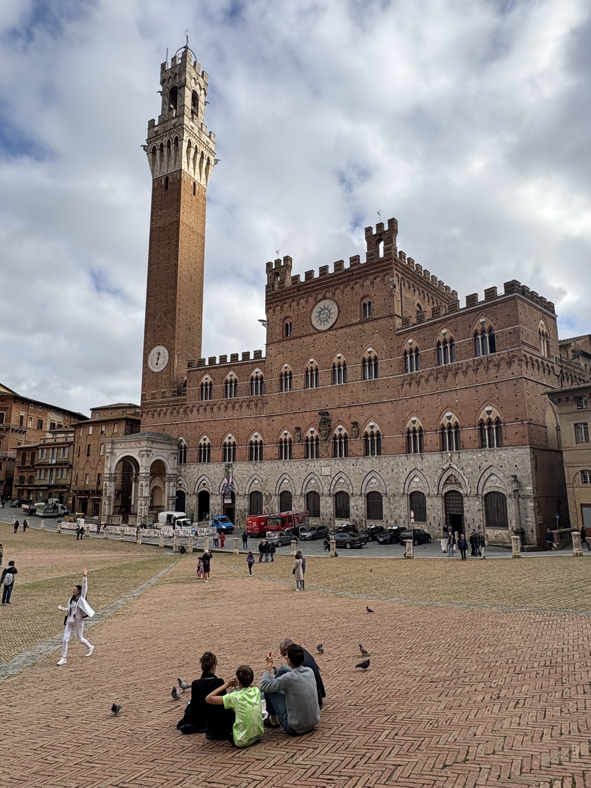 People sitting on a brick plaza in front of a historic red brick building with a tall tower and clock faces.
