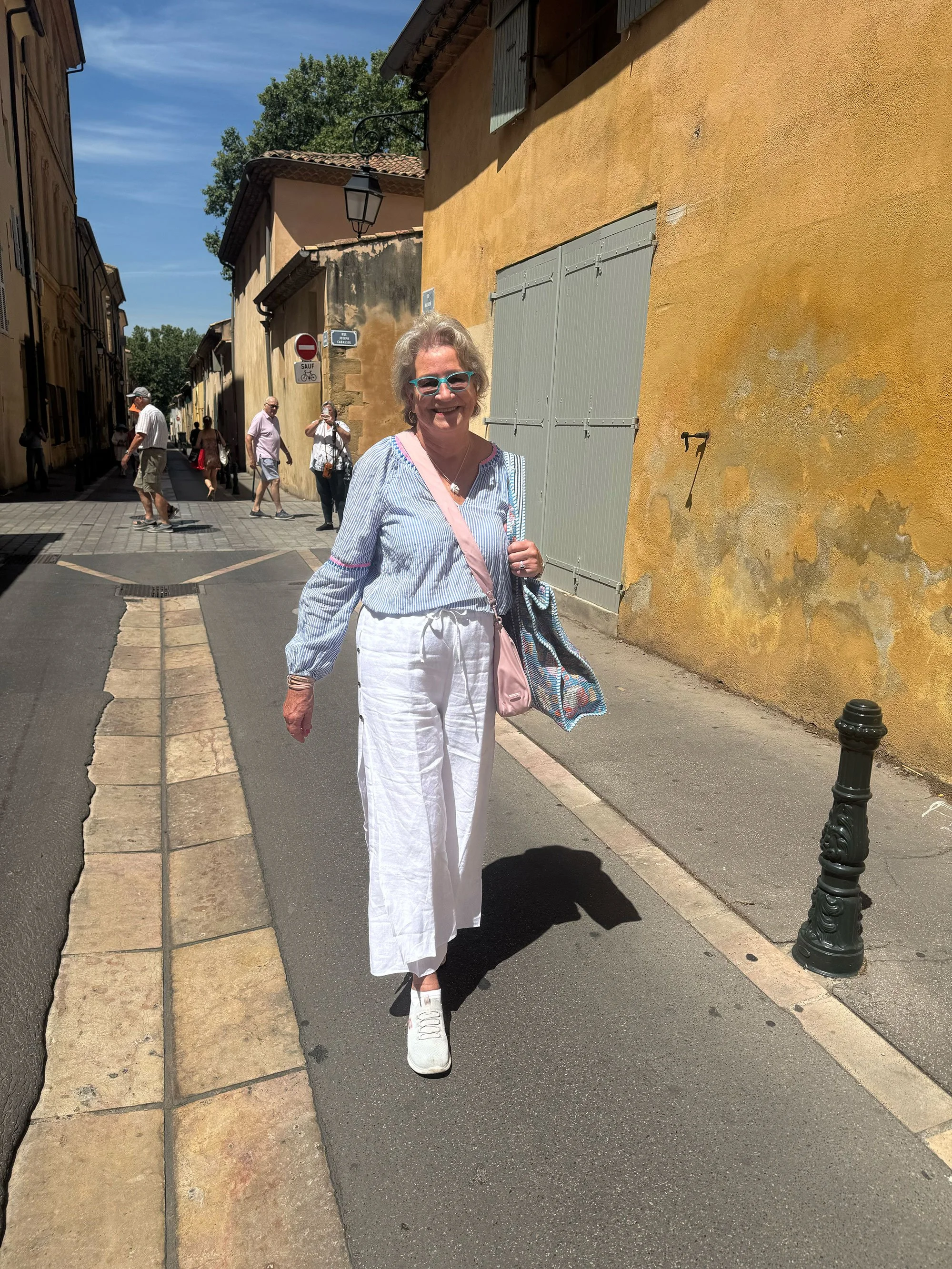 A woman walking on a cobblestone sidewalk in front of a yellow wall on a sunny day, wearing white pants, a striped blue and white top, white sneakers, sunglasses, and carrying a colorful bag.