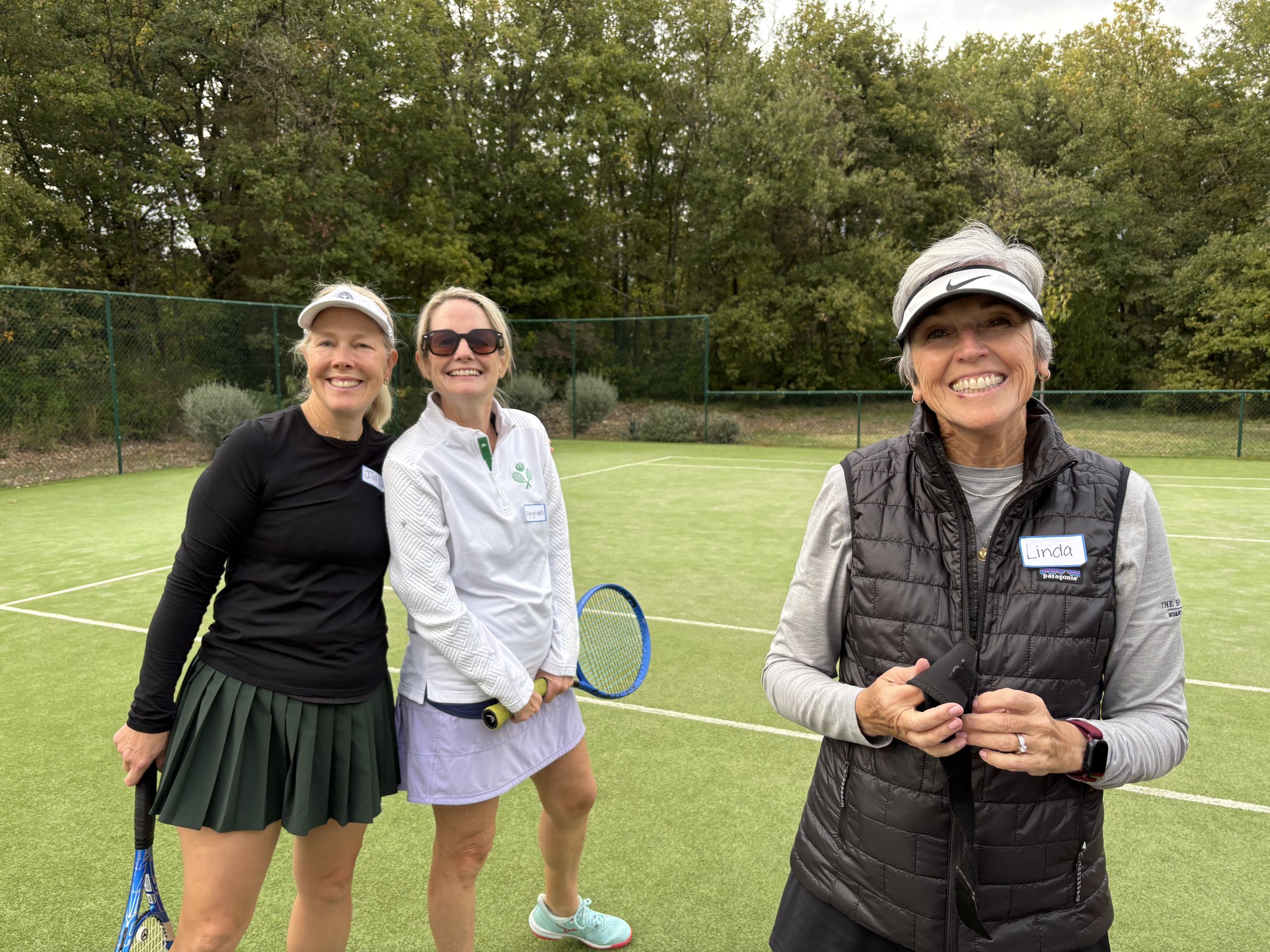 Three women standing on a tennis court outdoors, smiling. One is holding a tennis racket. The background shows trees and a green fence.