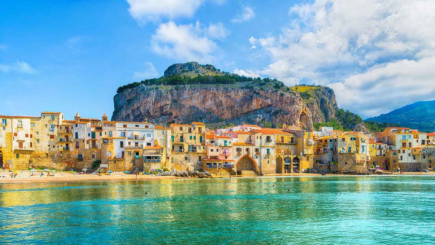 Colorful coastal village with buildings along the water, a large rocky hill behind, and a partly cloudy sky in Verdura, Taormina & Palermo.