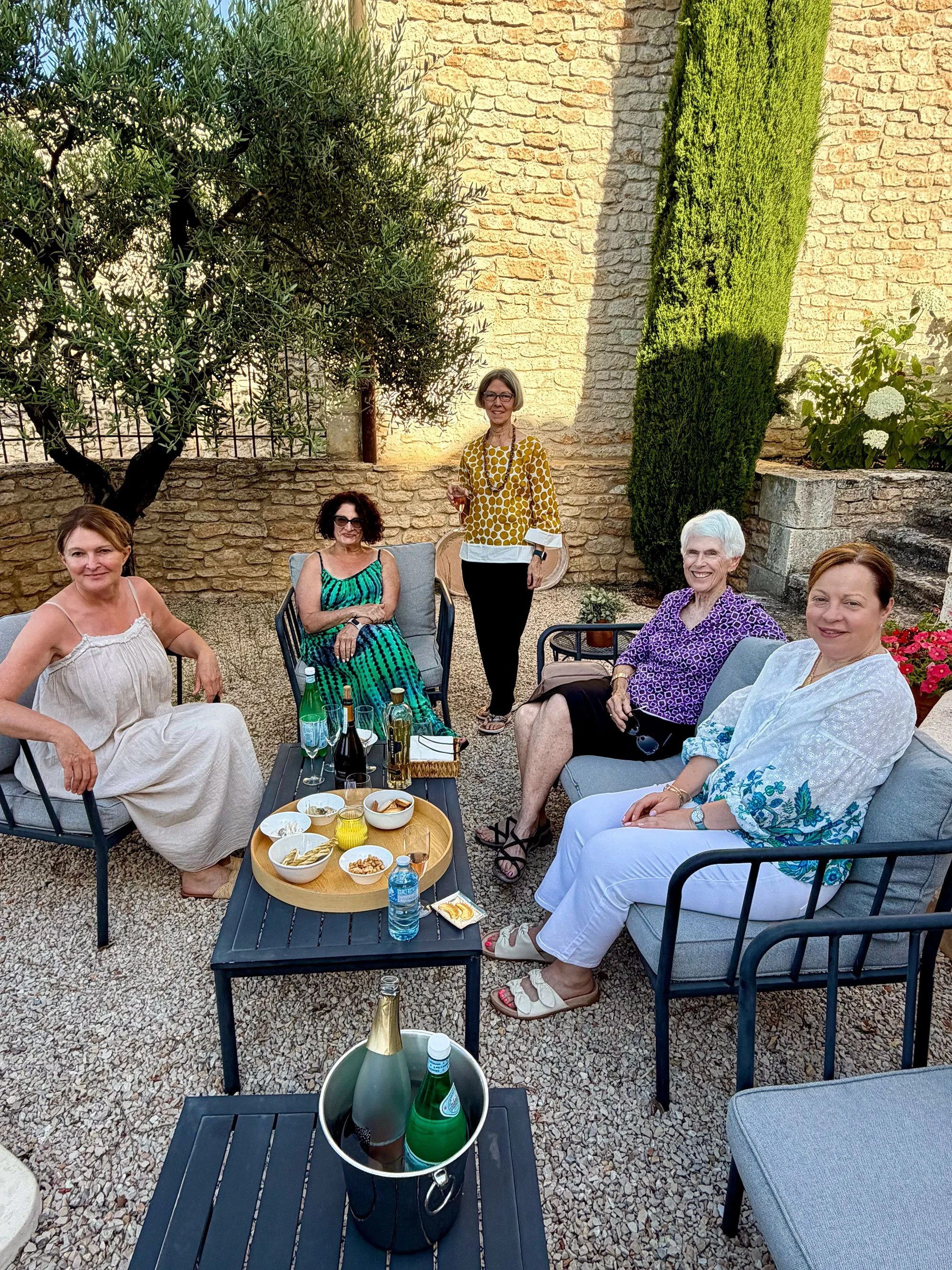 A group of five women sitting and standing outdoors, smiling and enjoying drinks in a garden with a stone wall, green shrubbery, and trees. There is a black table with snacks and drinks, including wine and sparkling water, in front of them.