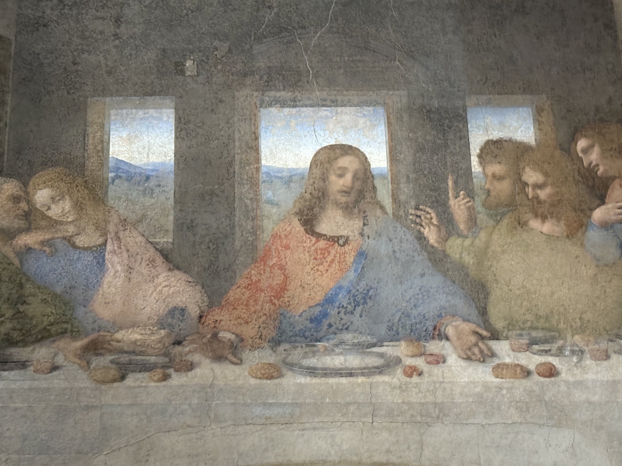Painting of the Last Supper showing Jesus Christ and his disciples at a long table with bread and goblets inside a room with three windows revealing mountains behind.