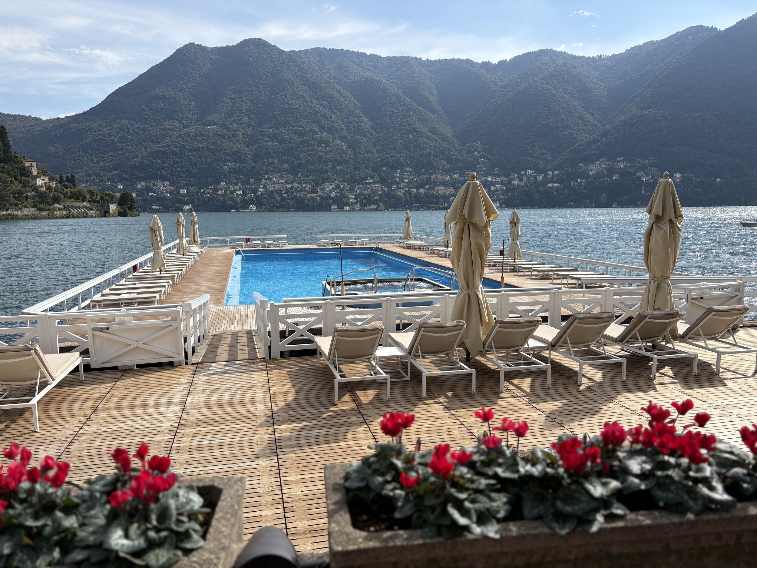 A lakeside outdoor swimming pool area with pool chairs and umbrellas, overlooking a mountain lake with mountain peaks in the background.
