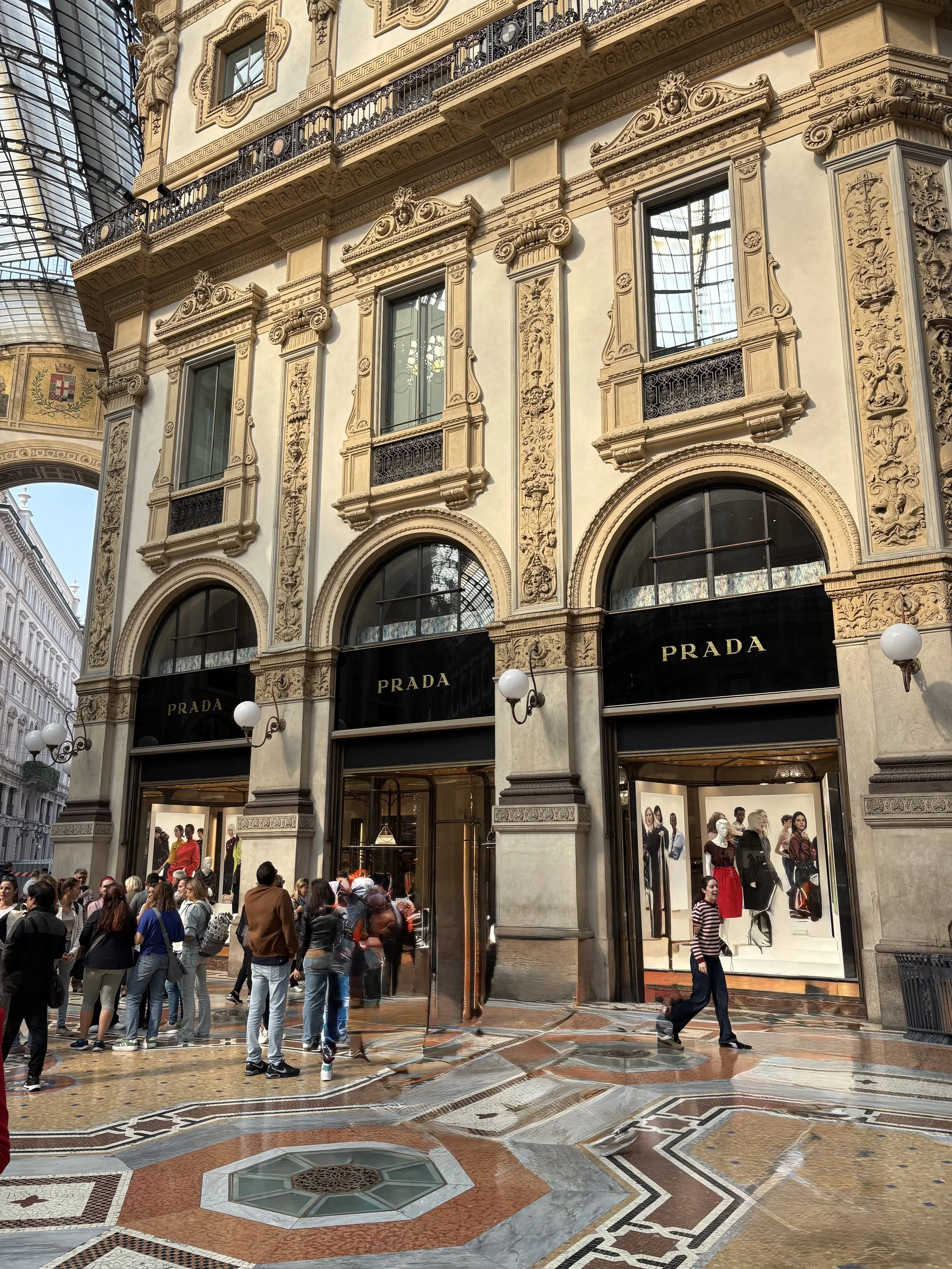 Exterior of a luxury Prada store in a historic building with ornate architectural details, located in a shopping arcade with a glass ceiling, and pedestrians in front.