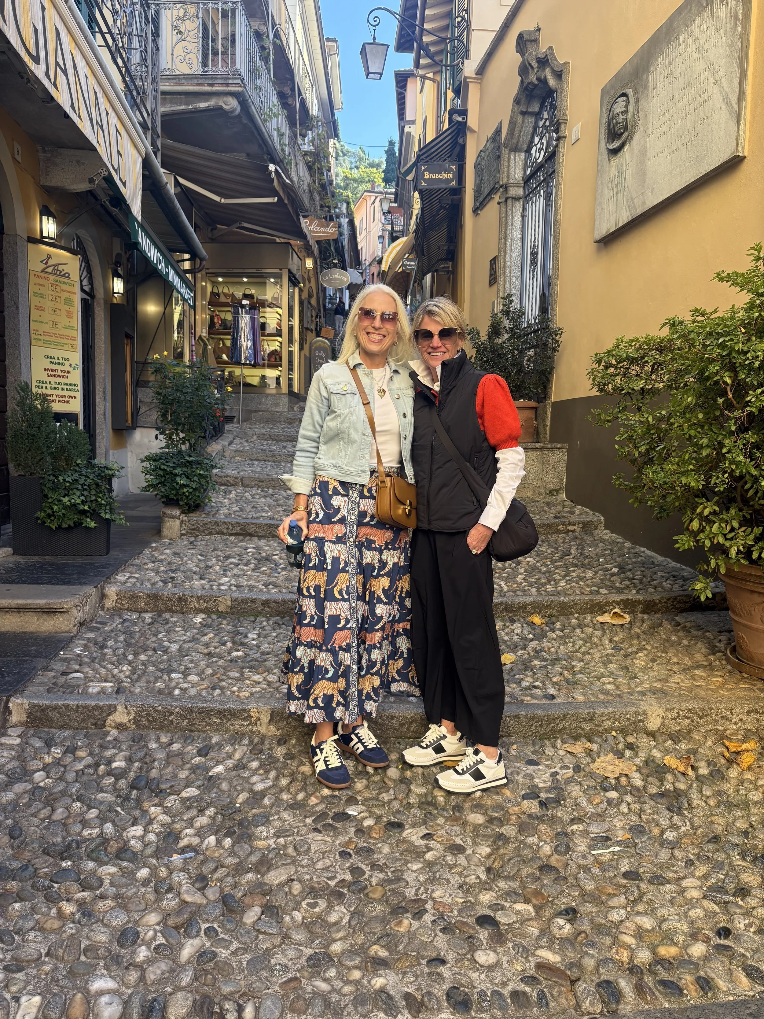 Two women standing on a cobblestone street in a European city, smiling, wearing sunglasses. One woman has long blonde hair, a light denim jacket, a colorful skirt, and sneakers. The other woman has short gray hair, a black vest with red sleeves, blac