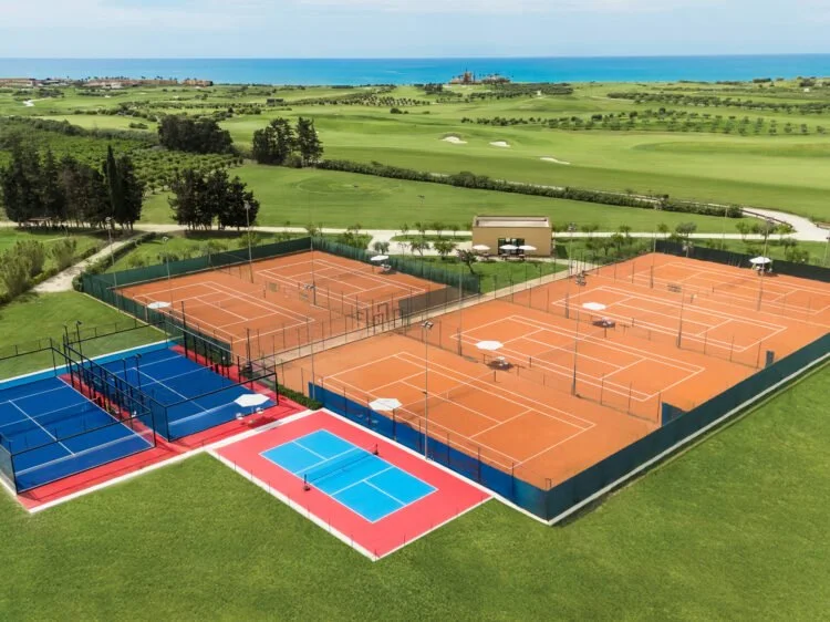 Aerial view of a sports complex with four tennis courts and one pickleball court, surrounded by green fields and a scenic ocean in the background.
