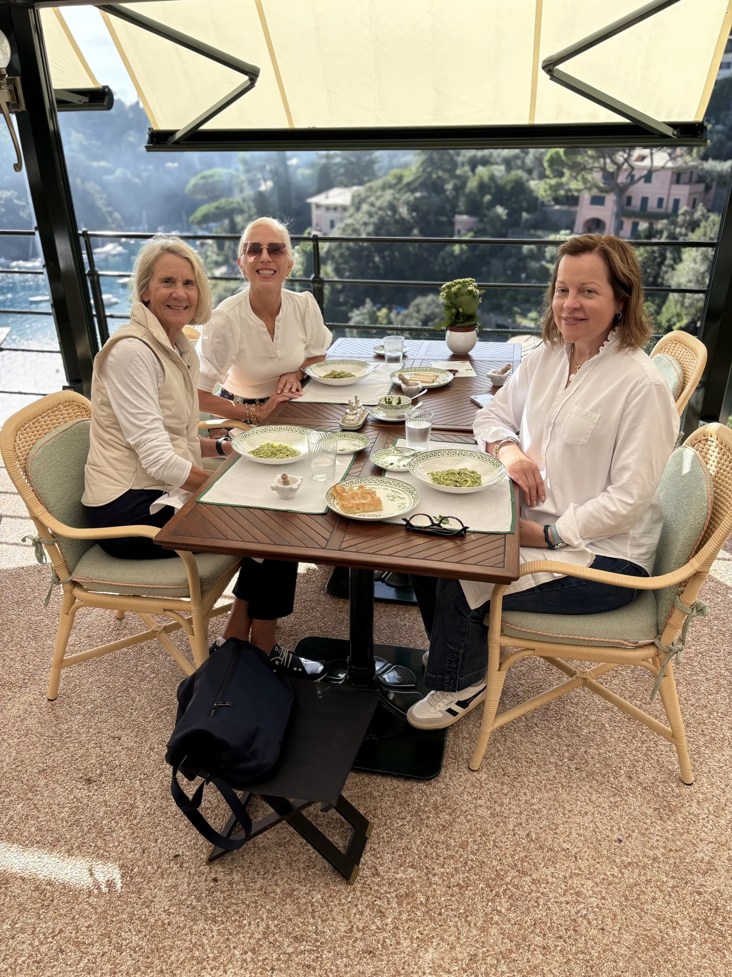 Three women sitting at a table with food on an outdoor terrace overlooking a hilly landscape with buildings and trees.