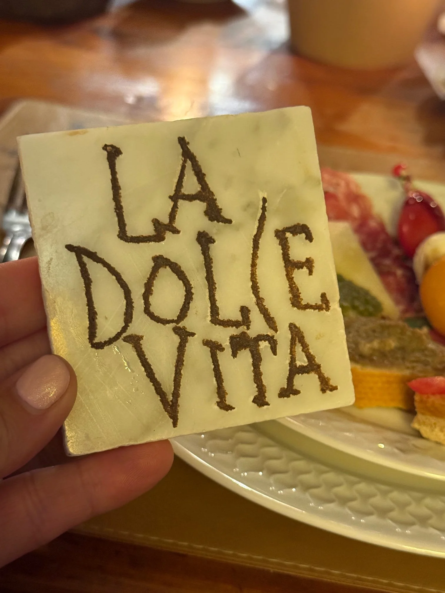 A hand holding a white square piece of cheese with the handwritten words 'LA DOLCE VITA' in dark substance. A plate of assorted foods is in the background.