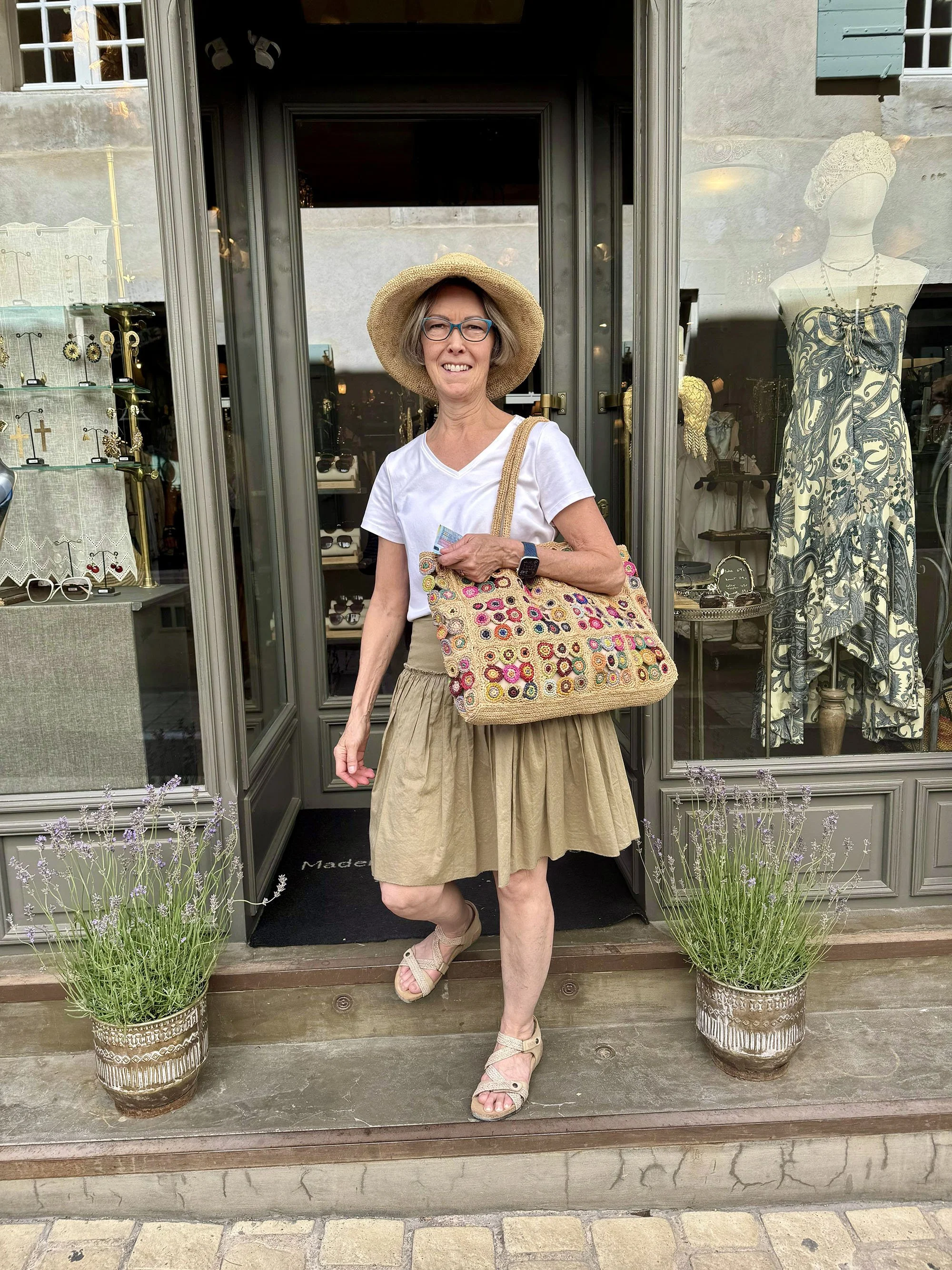 A woman stands in front of a boutique shop, wearing a straw hat, glasses, a white t-shirt, a beige skirt, and sandals, holding a colorful embroidered bag. The shop window displays jewelry and a dress mannequin.