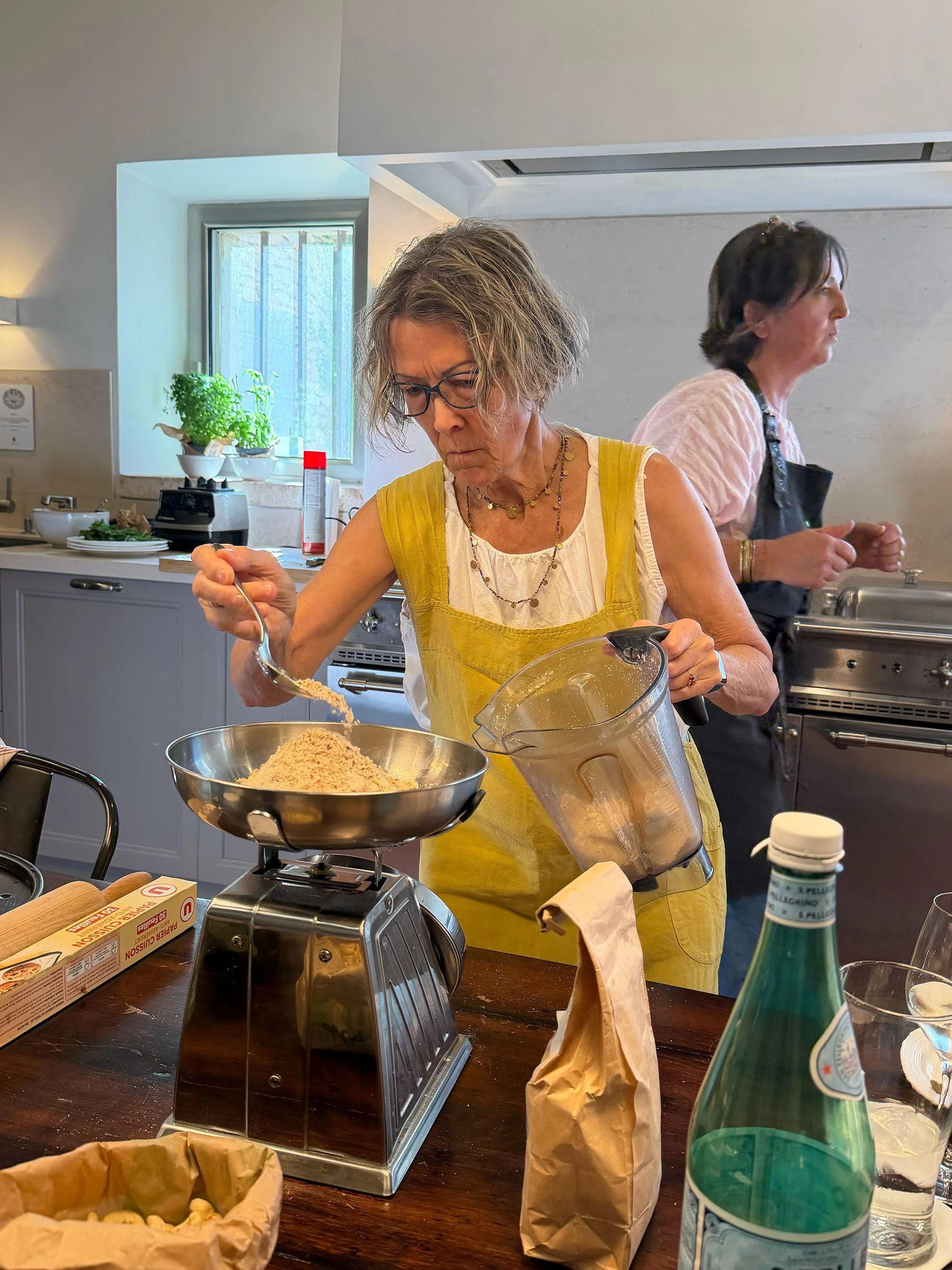 Two women are in a kitchen preparing food. The woman in the foreground with glasses and a yellow apron is pouring crumbs into a bowl, while the woman in the background is turned sideways, focused on her task. The countertop has a blender, a carton, a
