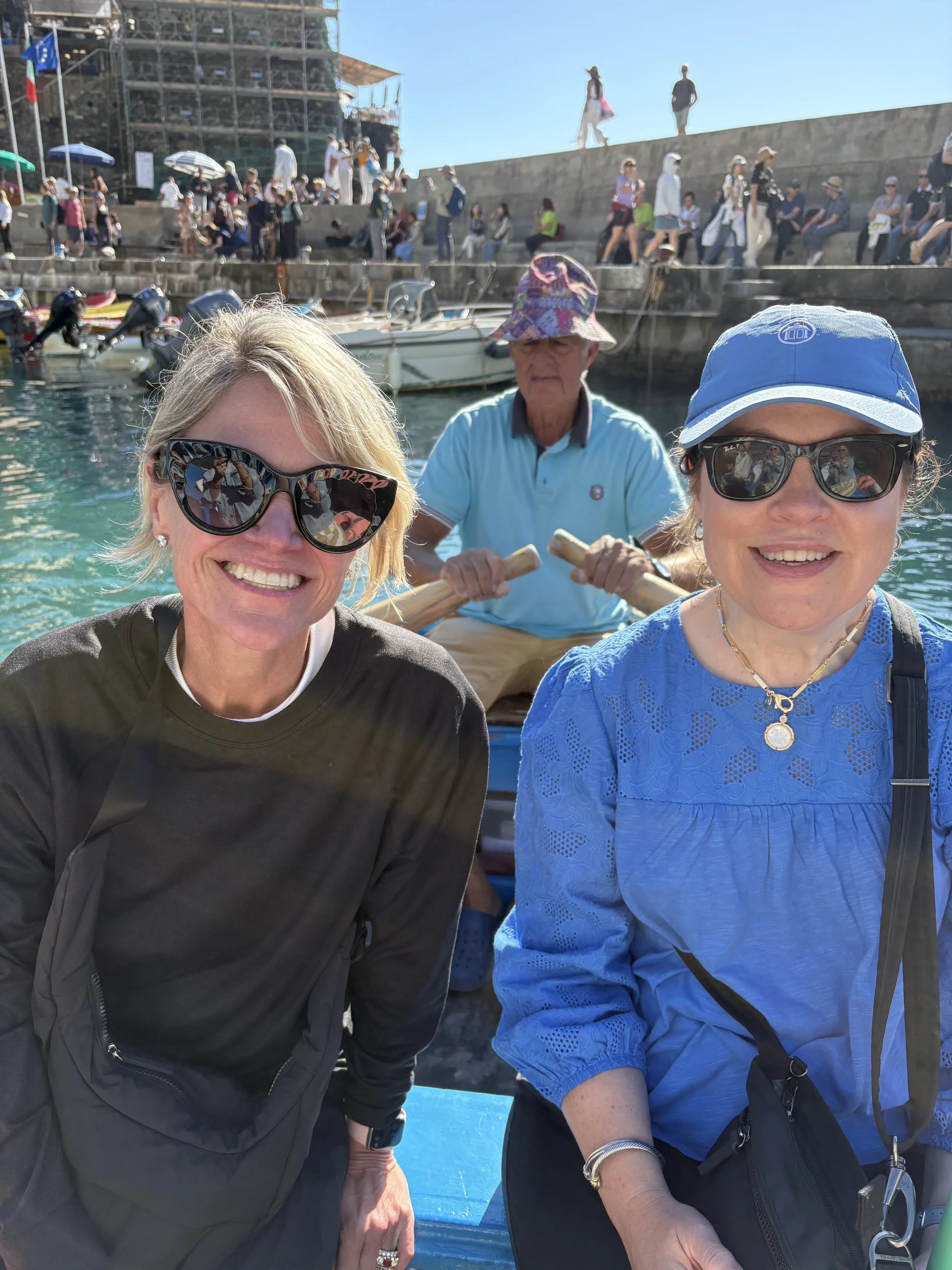 Two women smiling in the foreground, posing for a photo on a boat, with a man paddling behind them and a busy dock with people and boats in the background.