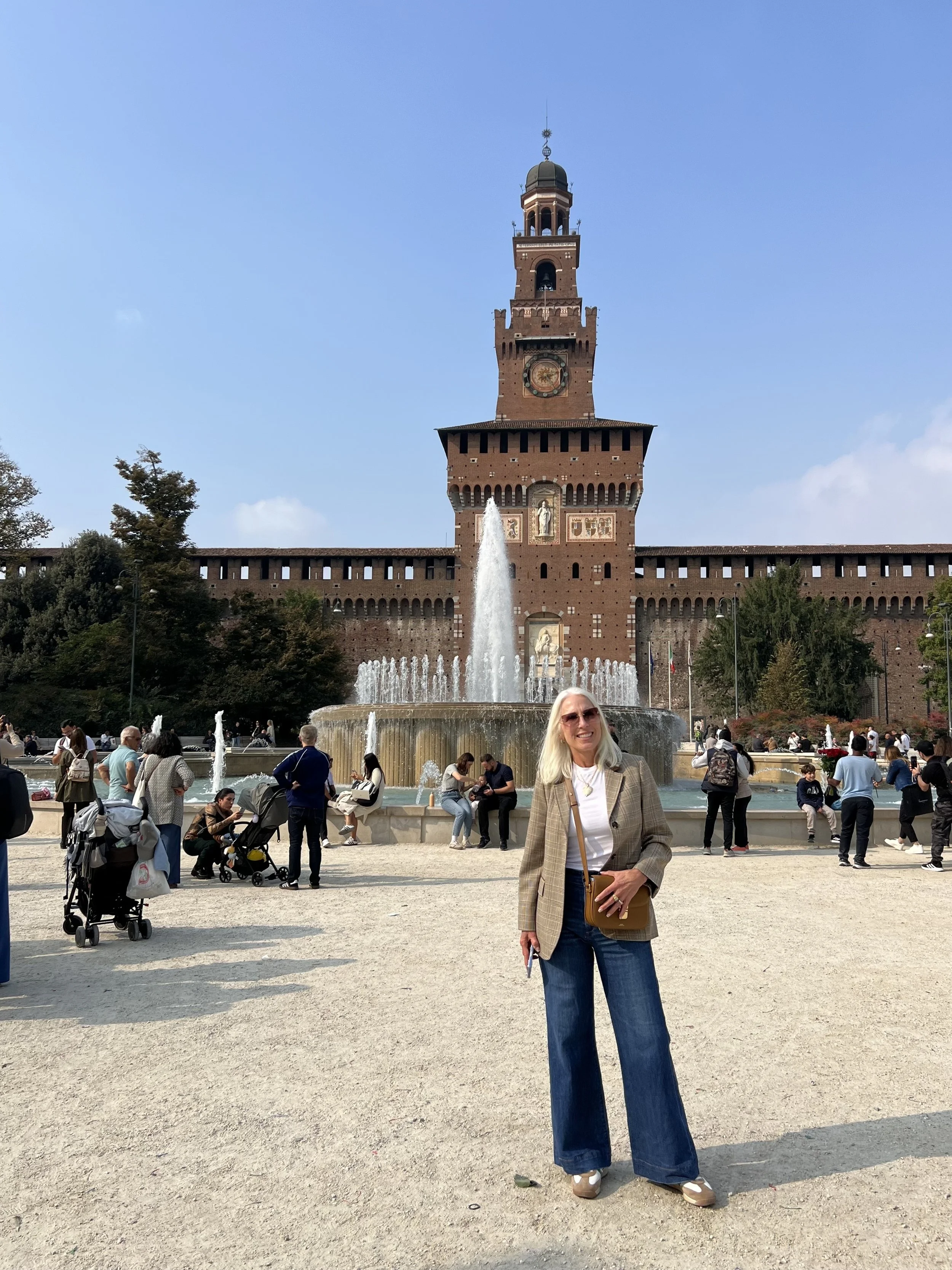 A woman with blonde hair wearing sunglasses, a plaid blazer, a white t-shirt, and wide-leg jeans, smiling and posing in front of a large fountain with a historic brick tower in the background, on a sunny day with a crowd of people around.
