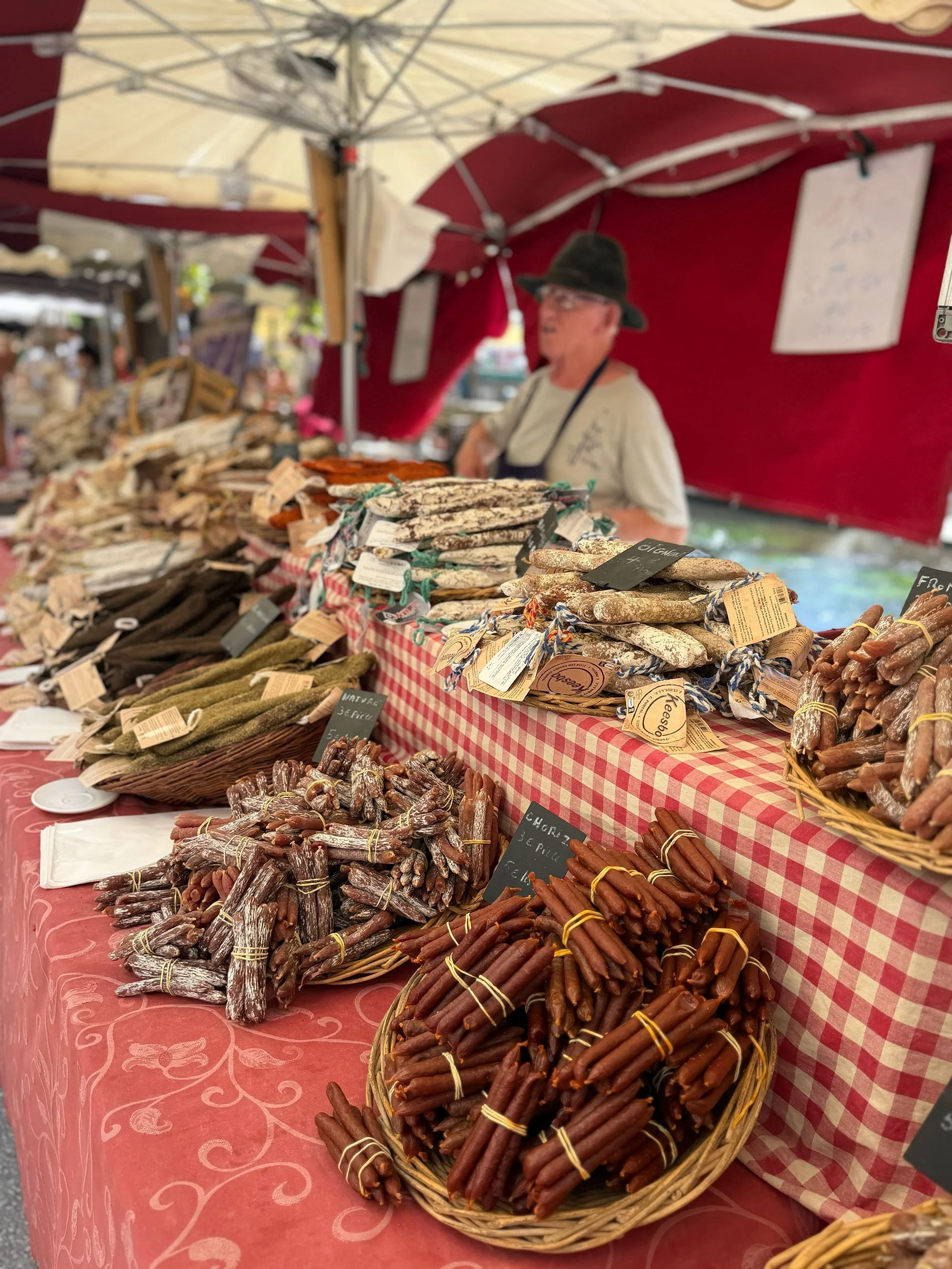 Baskets of assorted cured sausages and meats at a market stall with a red checkered tablecloth and a red canopy.