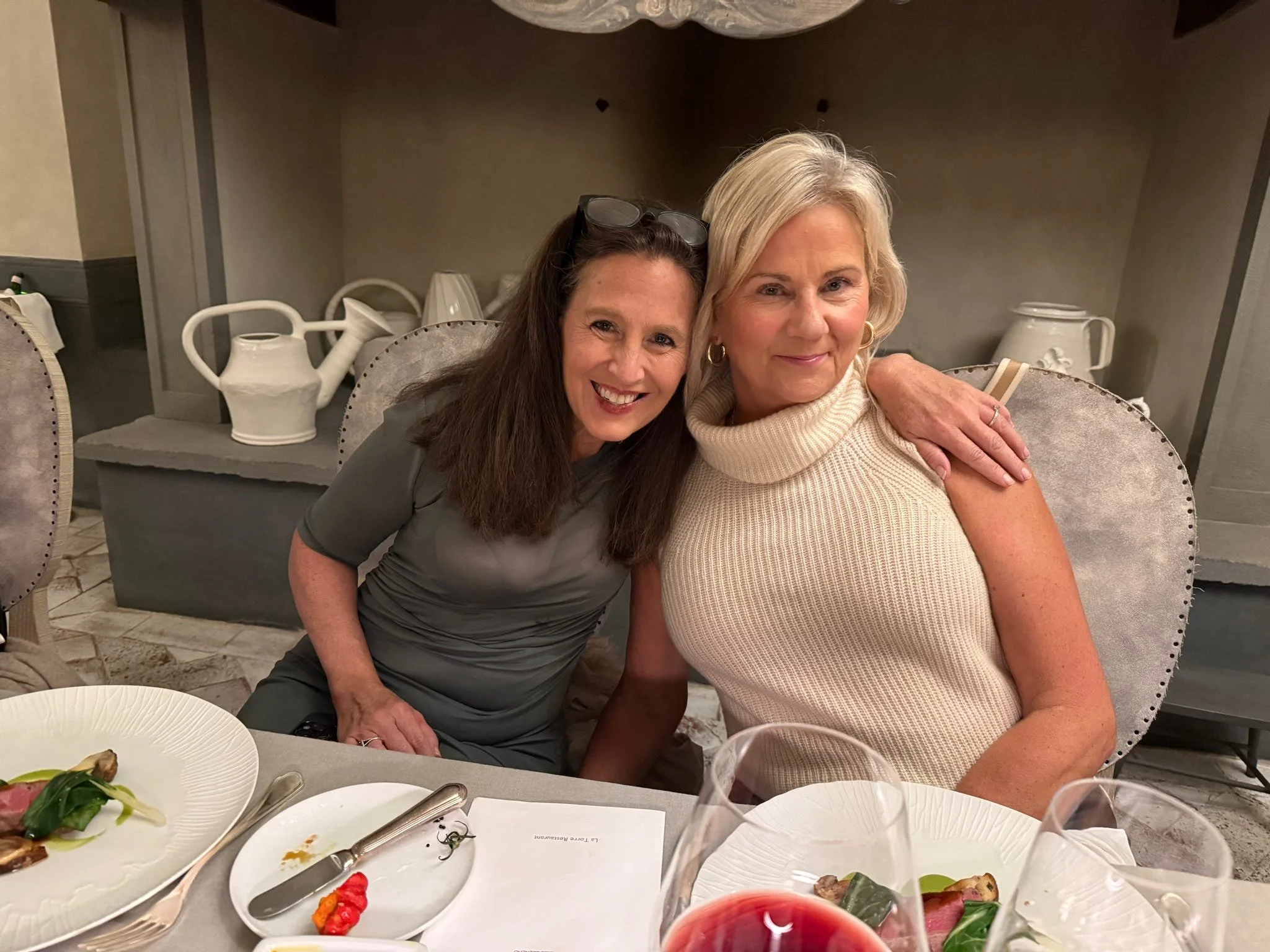 Two women sitting at a dining table, smiling at the camera. One woman has long brown hair, wearing glasses on her head and a gray dress. The other has short blonde hair, wearing a cream turtleneck top. The table has plates of food, wine glasses, and 
