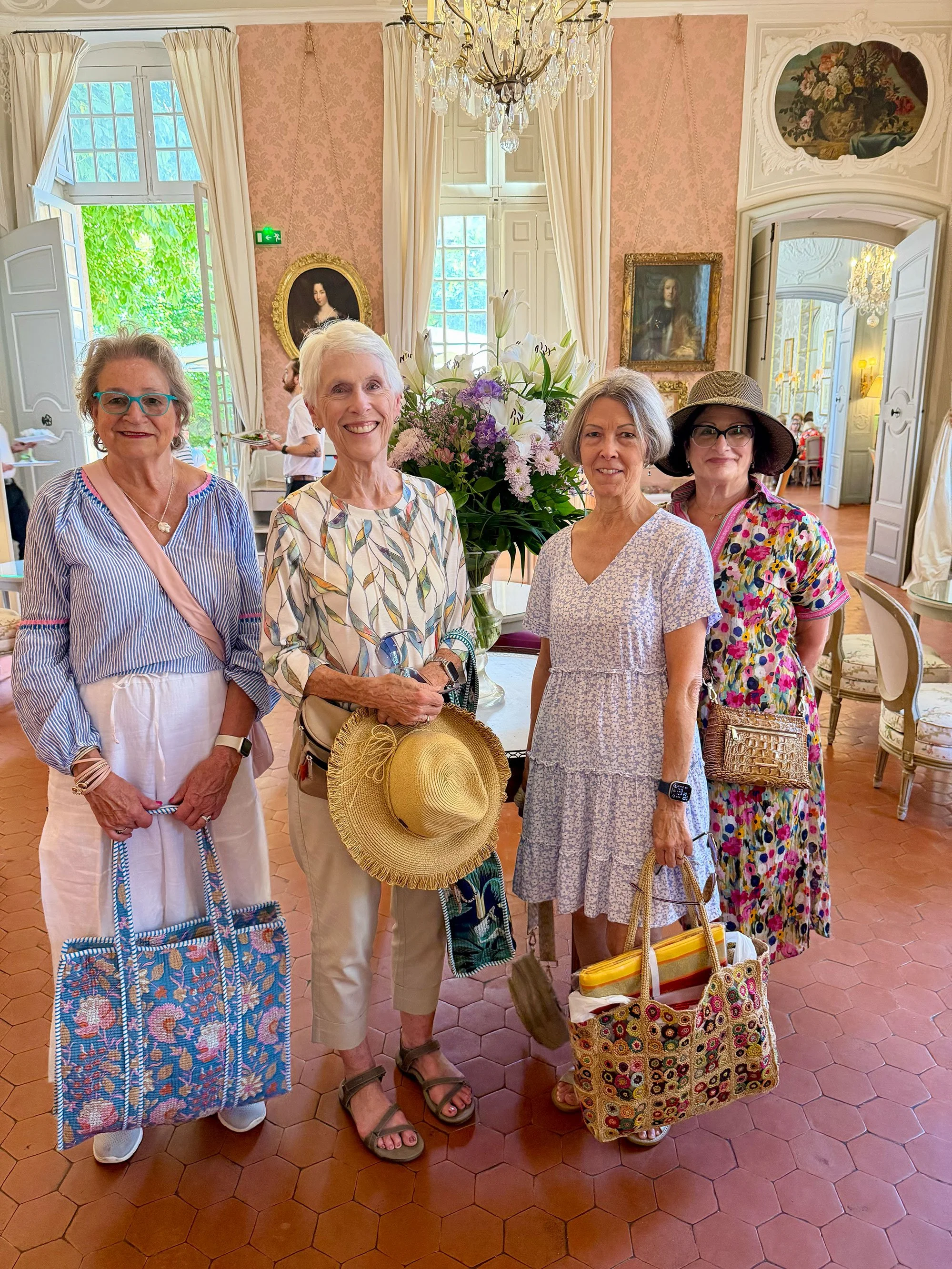 Four women standing in a luxurious, vintage-style room with pink damask wallpaper, large windows with white curtains, and a chandelier. They are smiling and holding handbags, with one woman also holding a wide-brimmed hat. In the background, a large 