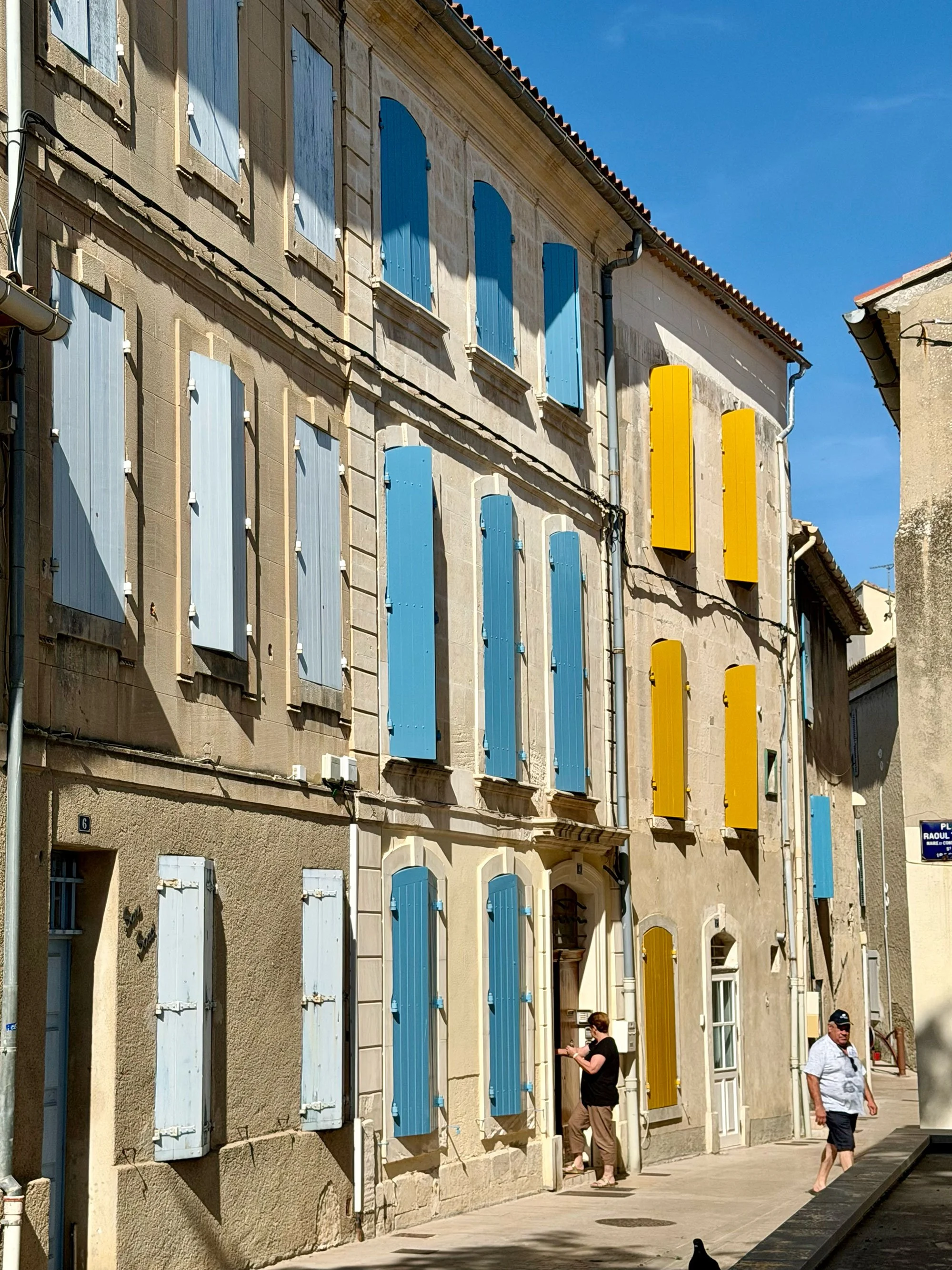 A street view of a building with beige stone walls and blue and yellow wooden shutters on the windows. There are two people walking near the building, one person is using a phone and the other is looking to the side. The sky is clear with some clouds
