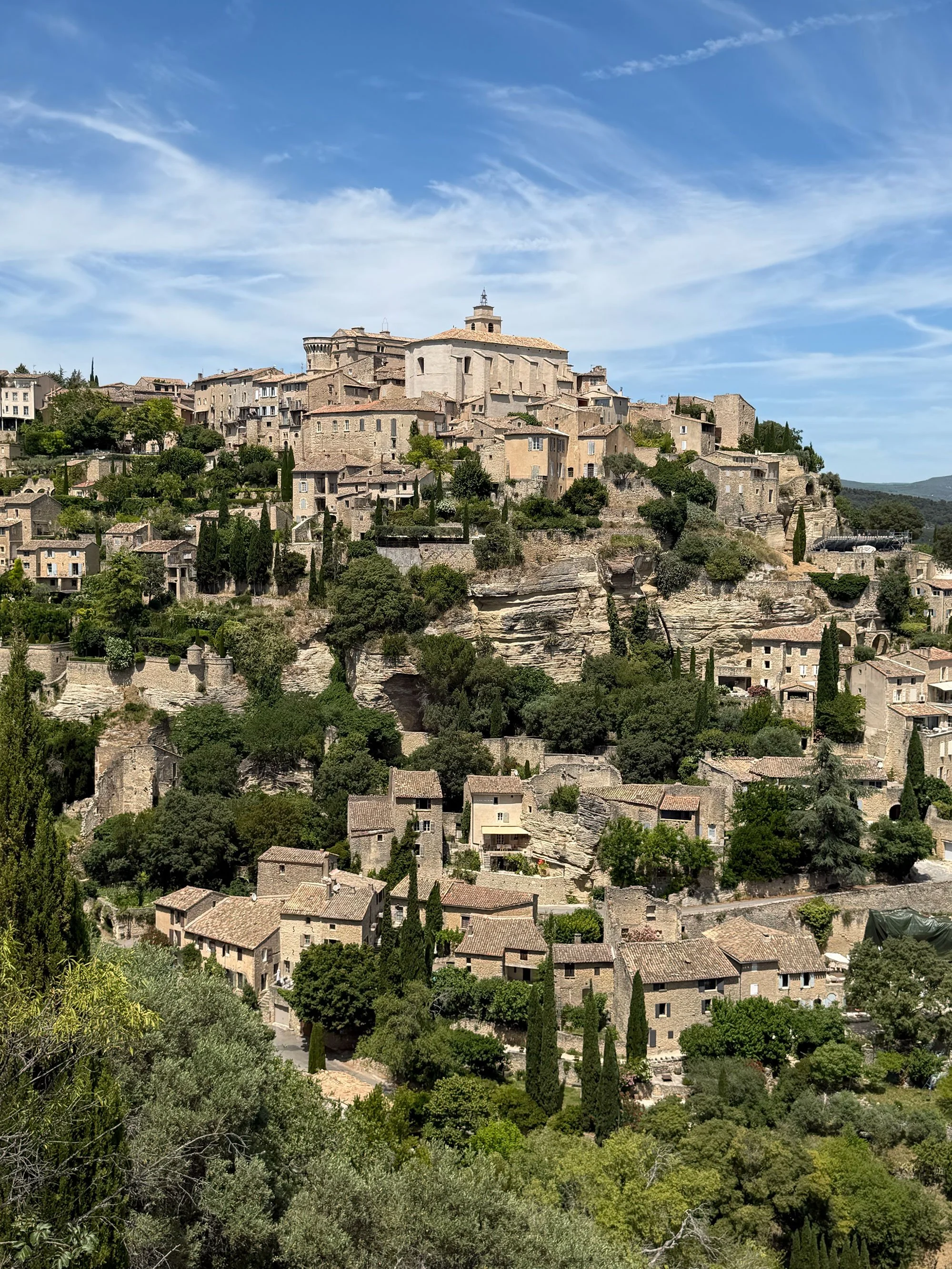 A historic hilltop village with stone buildings, a large central church, and lush green trees under a blue sky with wispy clouds.