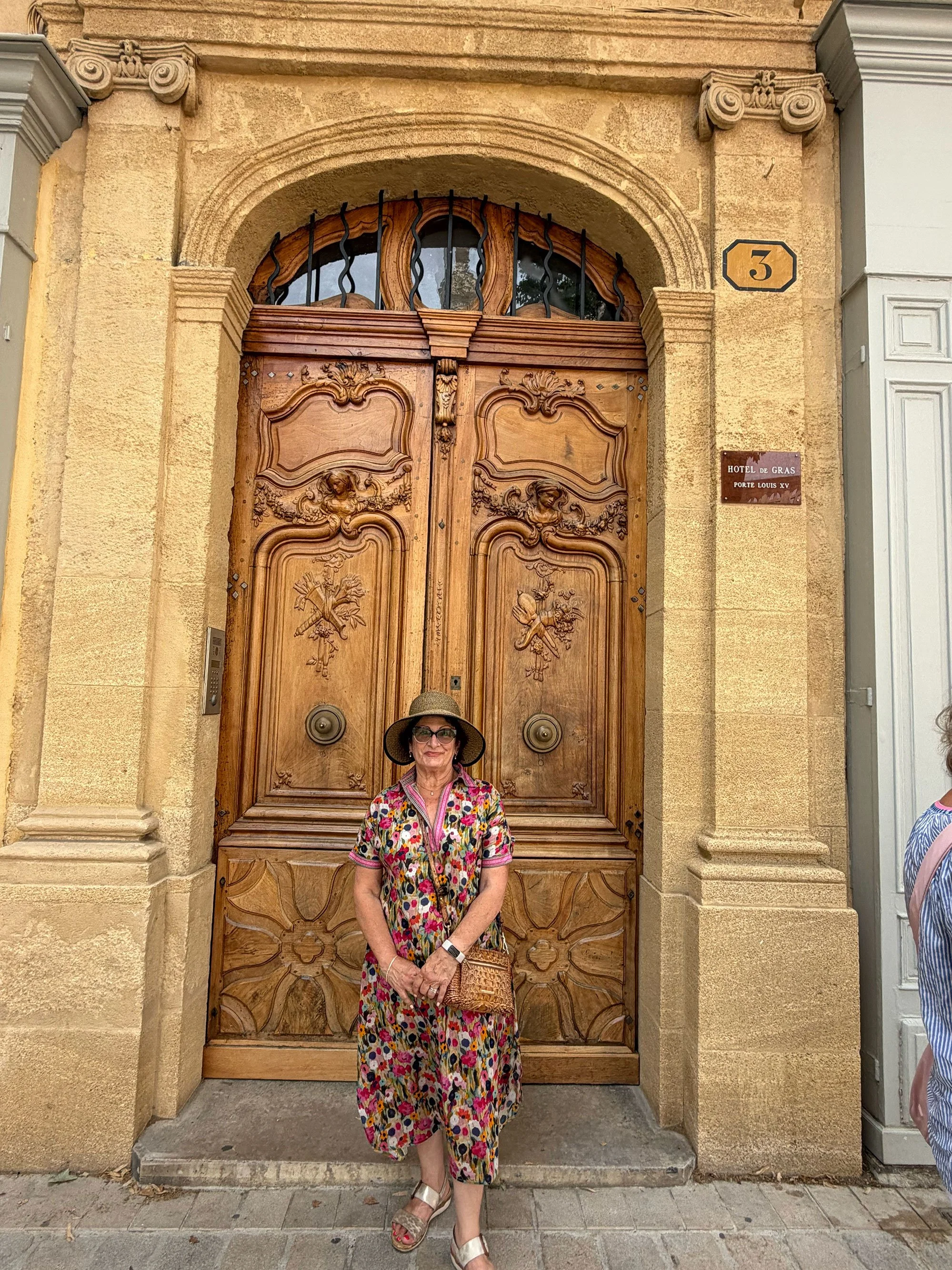 A woman standing in front of a large wooden door with ornate carvings, wearing sunglasses, a floral dress, a wide-brimmed hat, and carrying a woven handbag.