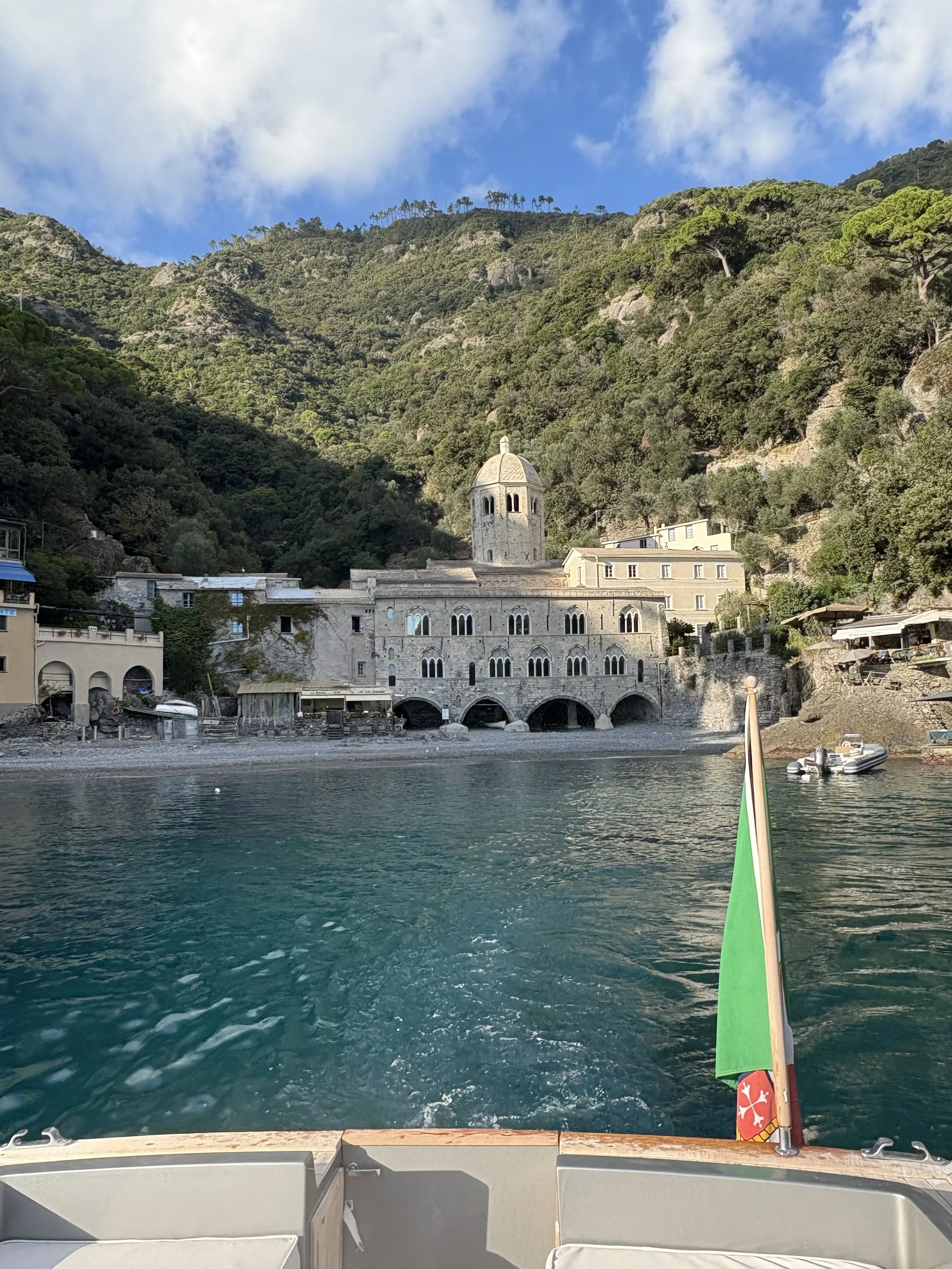 View from a boat on the water approaching a historic stone building and hillside covered with greenery in a coastal town.