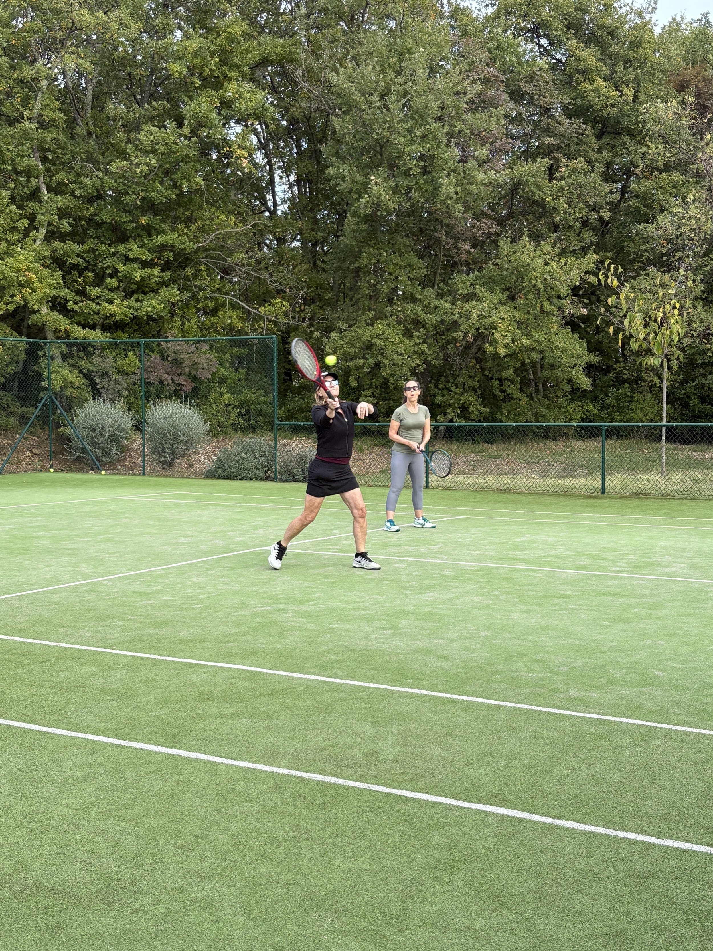Two women playing tennis on a green tennis court with trees in the background.