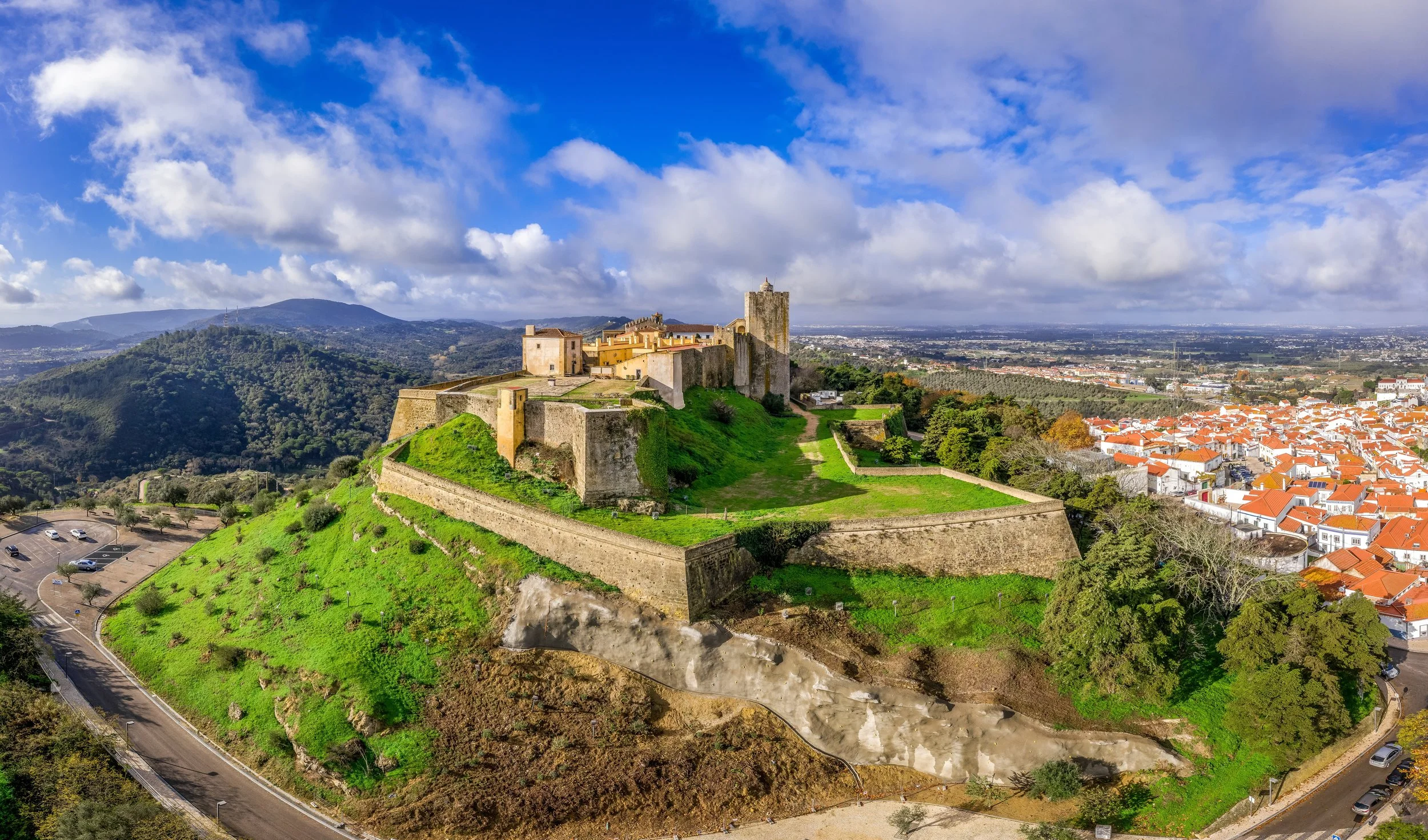 Aerial view of a historic hilltop fortress with stone walls and towers overlooking a town with white buildings and orange roofs, under a partly cloudy sky.