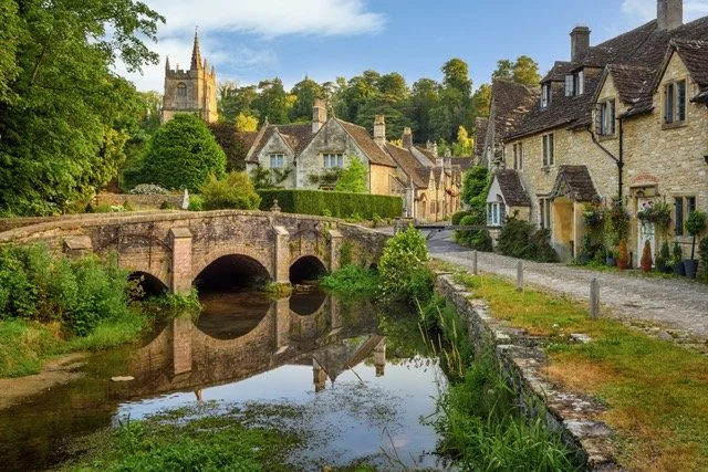 A picturesque village scene with a stone bridge over a calm river, historic stone houses lining a narrow street, lush greenery, and a church steeple in the background under a partly cloudy sky in Oxfordshire, Cotswolds & Cornwall.