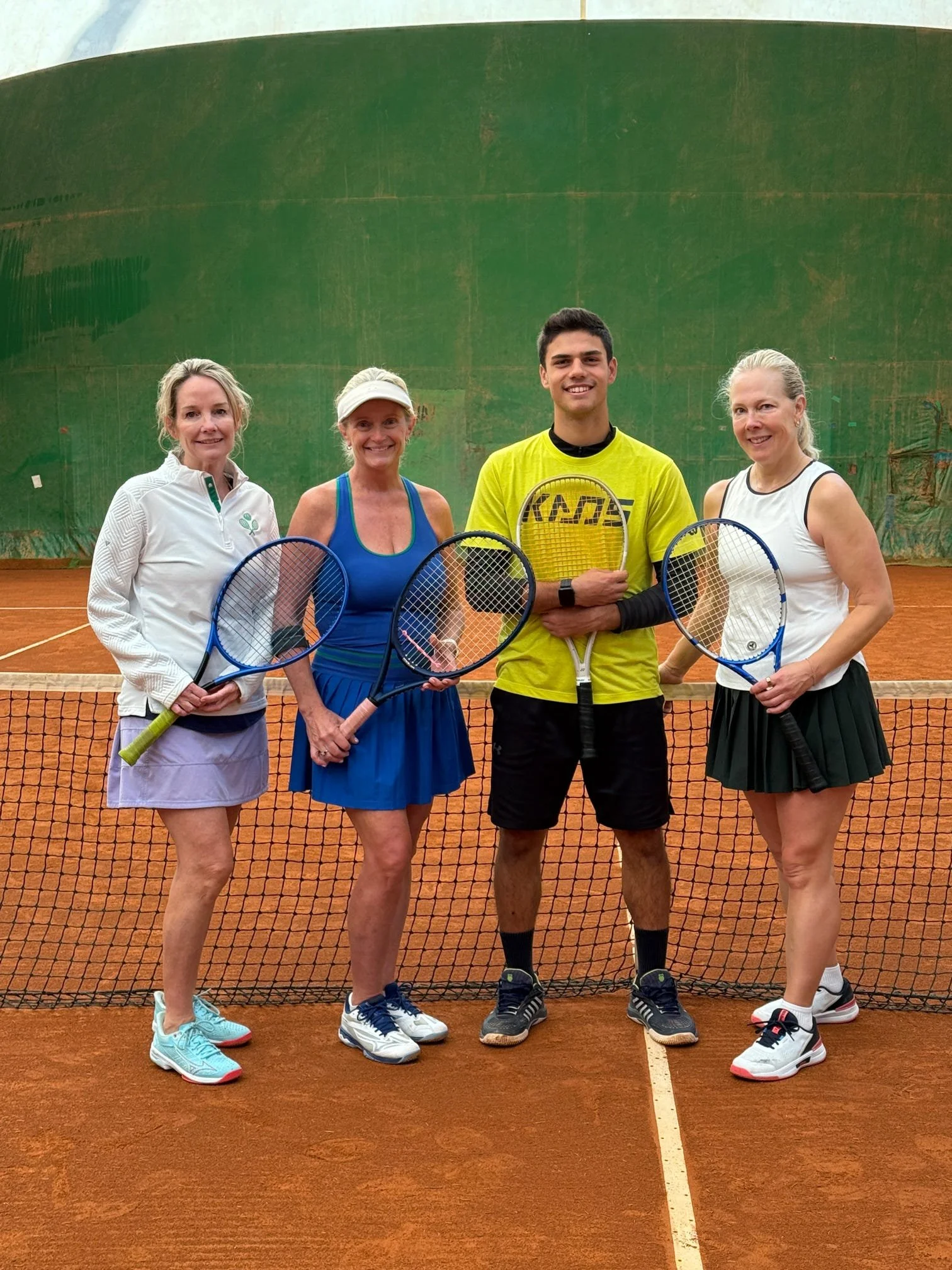 Four women and one young man standing on a clay tennis court, holding tennis rackets, with a green back wall behind them.