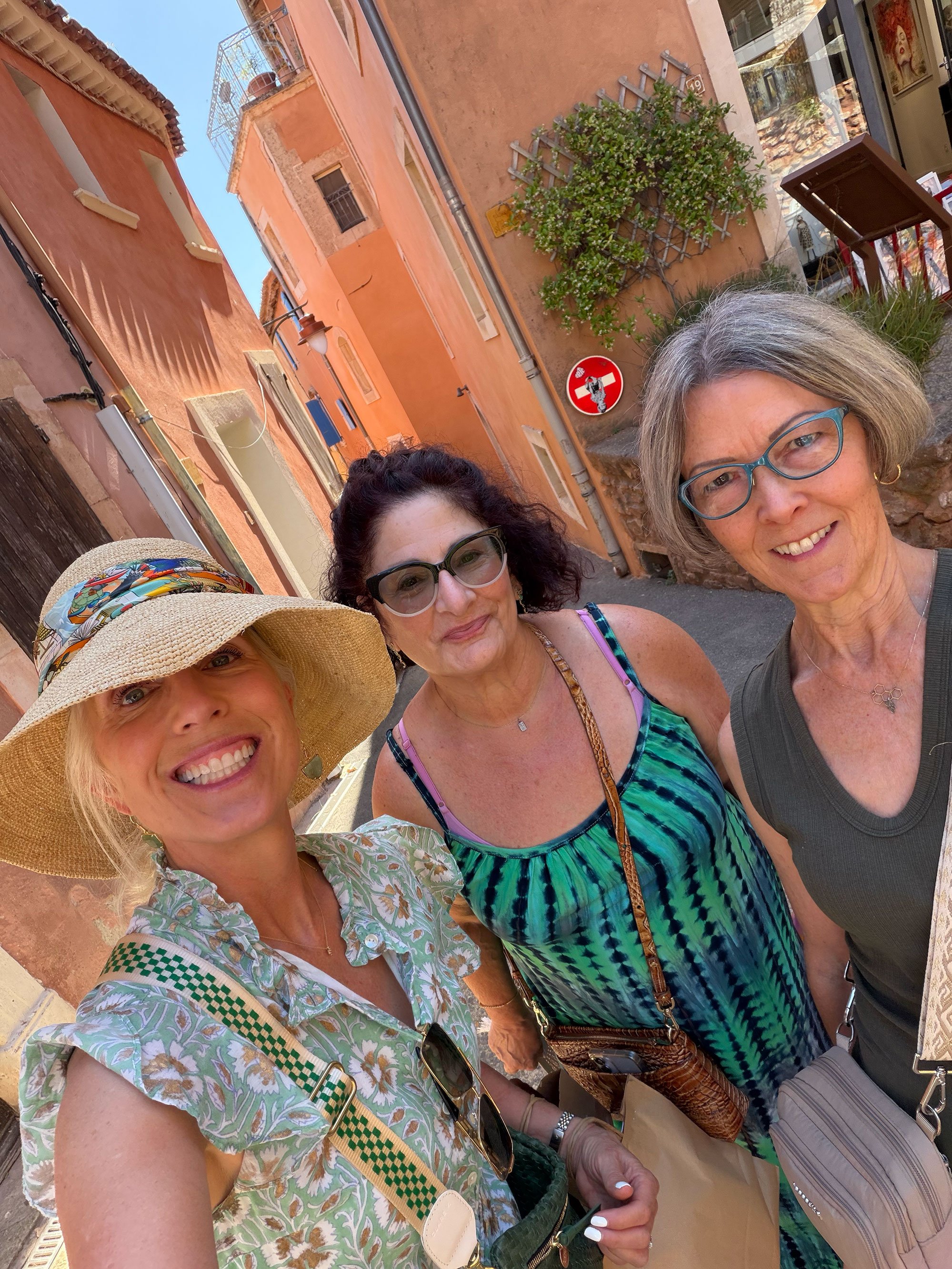 Four women taking a selfie together on a sunny street with colorful buildings and greenery in the background.