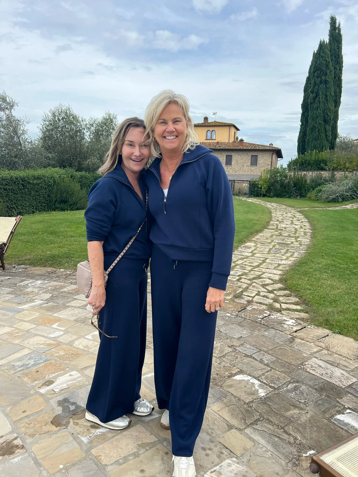 Two women wearing matching blue tracksuits smiling and standing close together outdoors on a stone-paved walkway with a curved stone path, lush green grass, bushes, and tall trees in the background during daytime.
