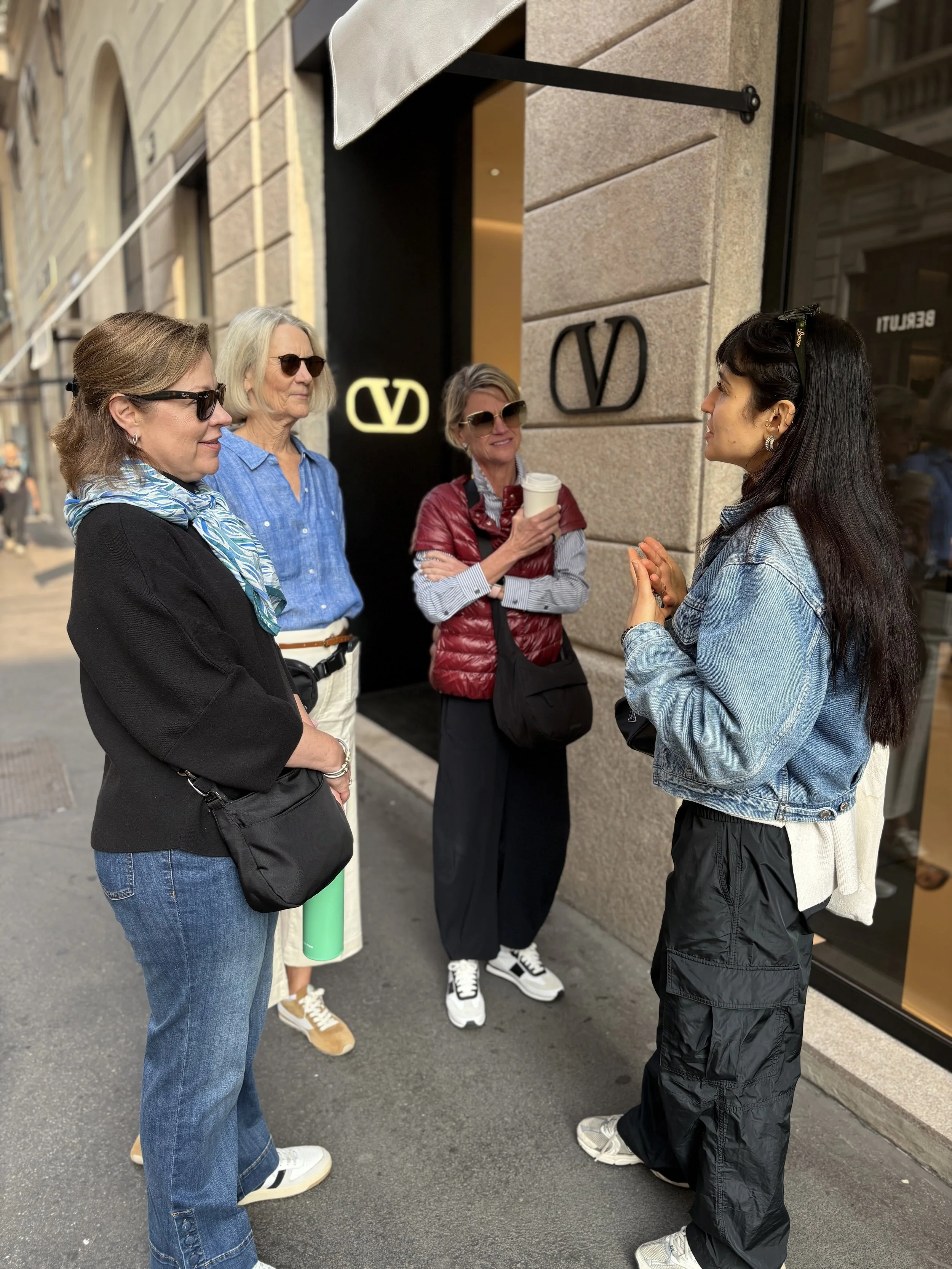 Four women having a conversation on a city sidewalk outside a store with a Valentino logo