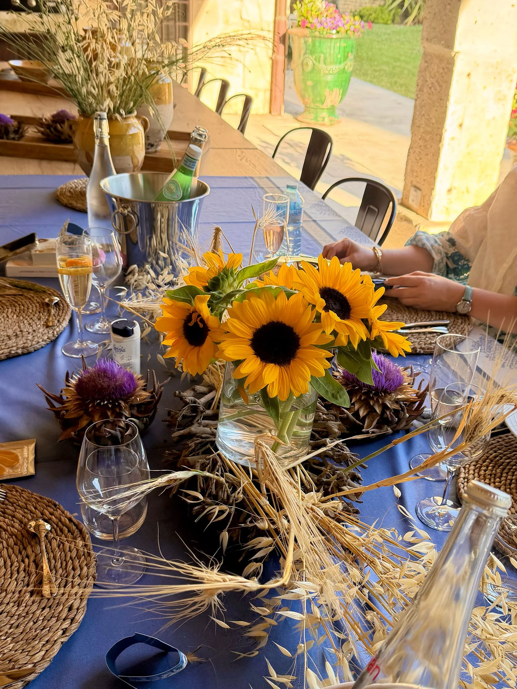 A dining table decorated with bright yellow sunflowers, purple thistles, and dried wheat and straw, set with wine glasses, a bottle of water, and a bottle of sparkling water, with woven placemats and a blue tablecloth, in a bright outdoor setting wit