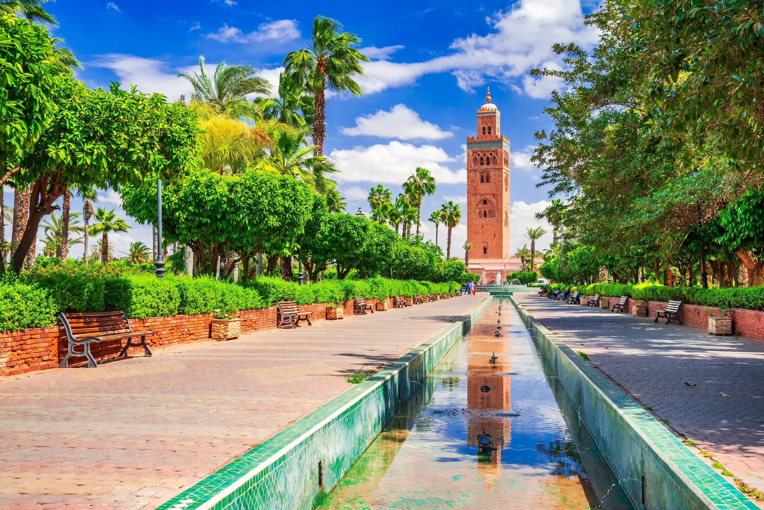 A scenic park with lush green trees lining a paved walkway and a water fountain in the middle. In the background, a tall red brick minaret stands against a partly cloudy blue sky.