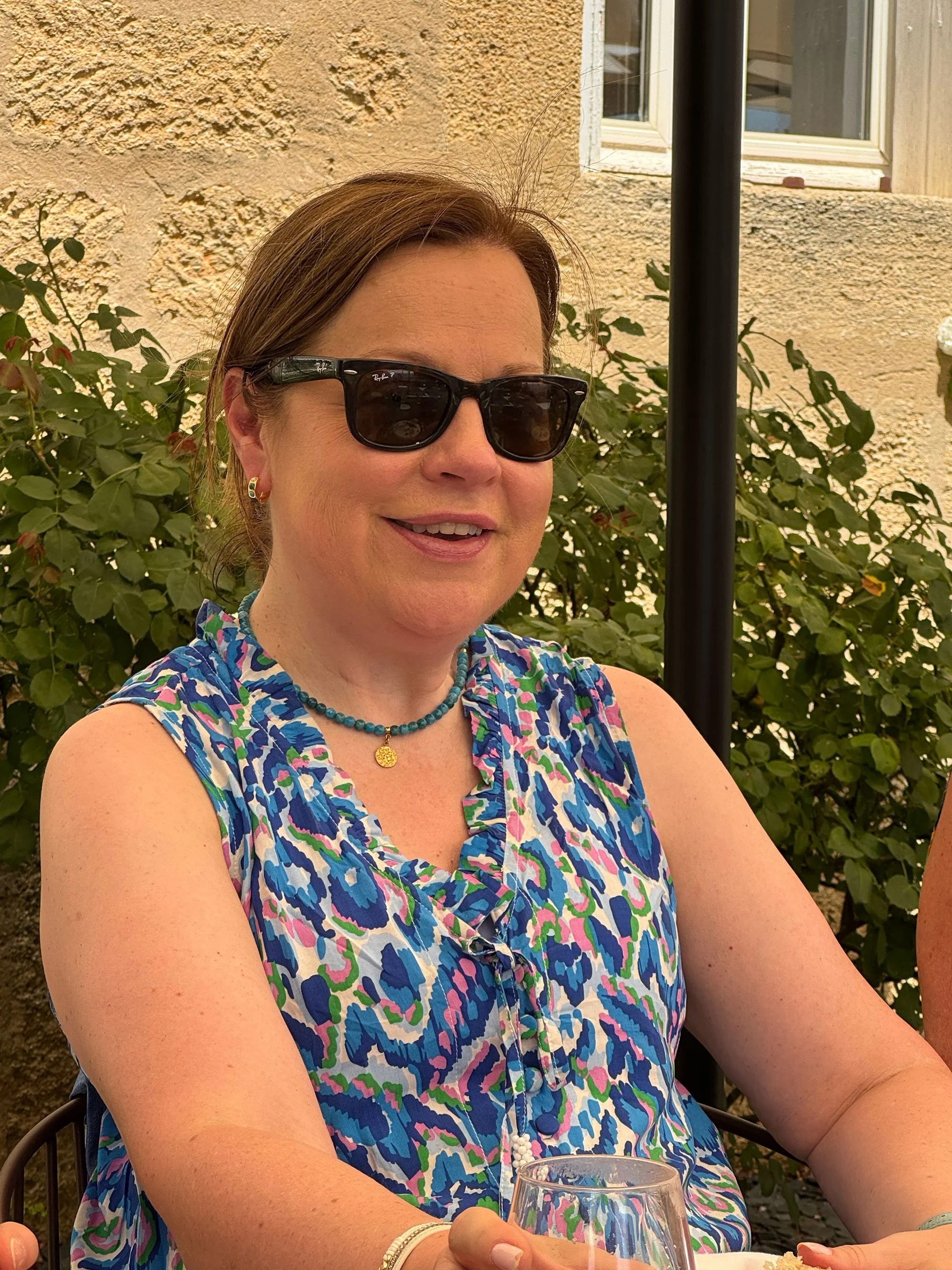 A woman wearing sunglasses and colorful sleeveless top sitting outdoors with a glass of wine, smiling, with a textured beige wall, window, and green shrubbery in the background.