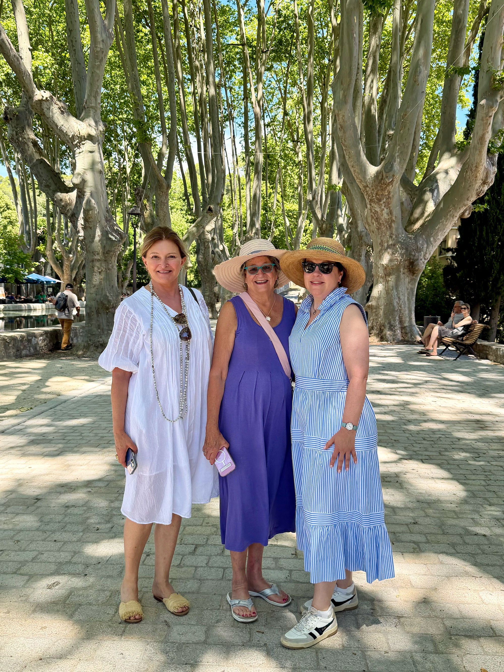 Three women standing together outdoors in front of large trees. They are dressed in summer clothing, wearing hats and sunglasses, and smiling at the camera. Other people are sitting on benches in the background.