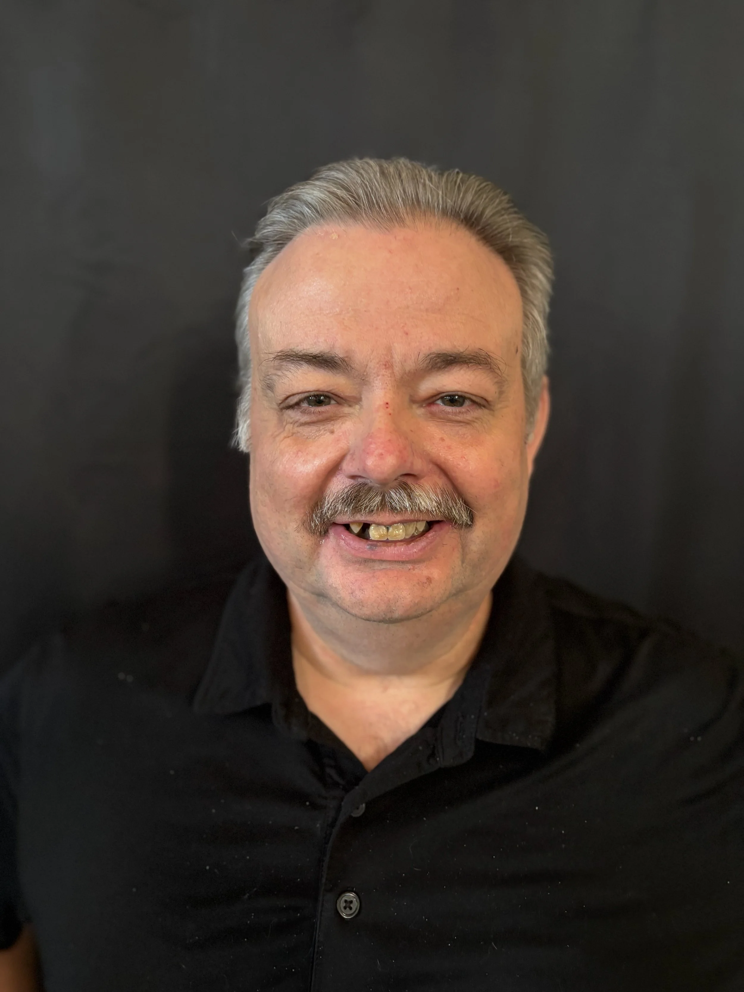 A smiling man with gray hair and a mustache standing outdoors with autumn trees in the background.