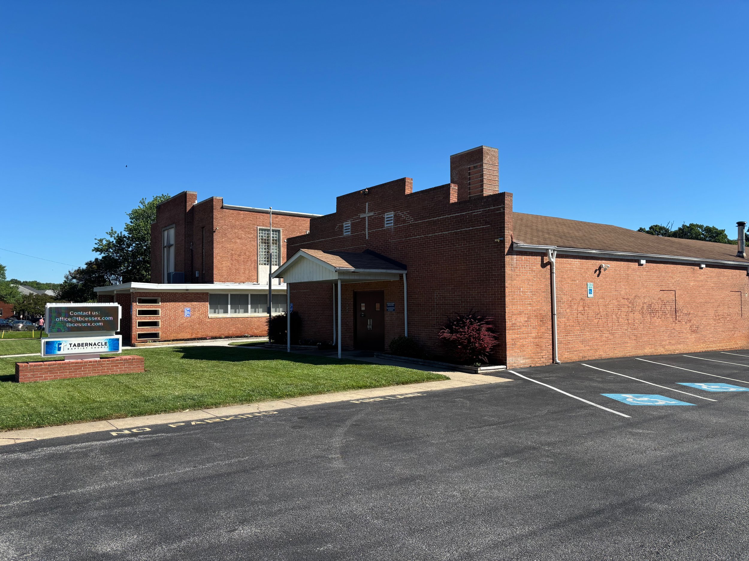 Exterior view of a church or community building with brick walls, a small covered porch, and nearby flower bed and grassy area, under a clear blue sky.