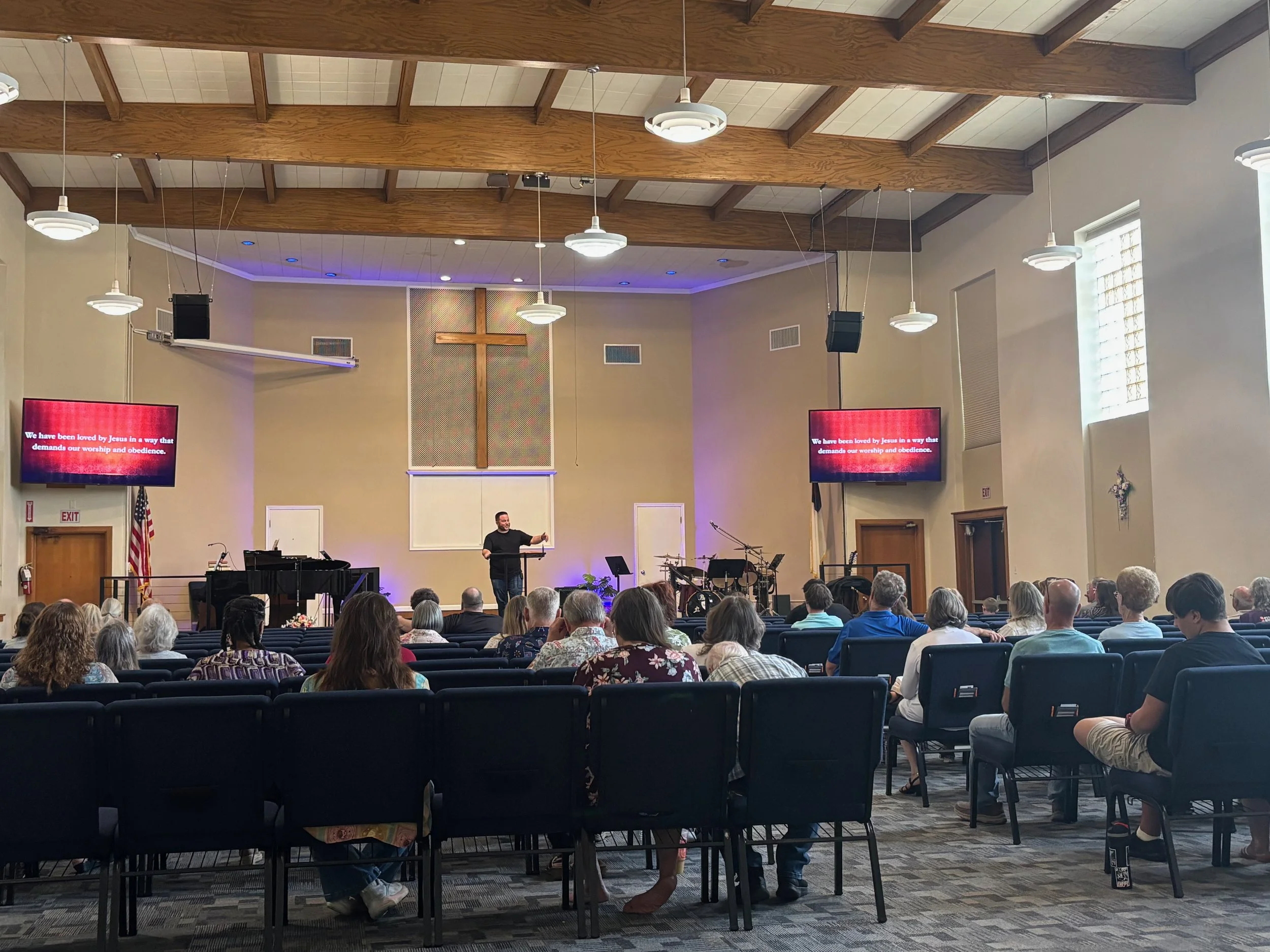 Inside a church with a high wooden vaulted ceiling, people seated in rows of chairs facing a stage where a man is speaking. The church has a large crucifix on the wall behind the stage, and two screens displaying religious messages. There are hanging lights, windows with blinds, and some stained glass decorations.