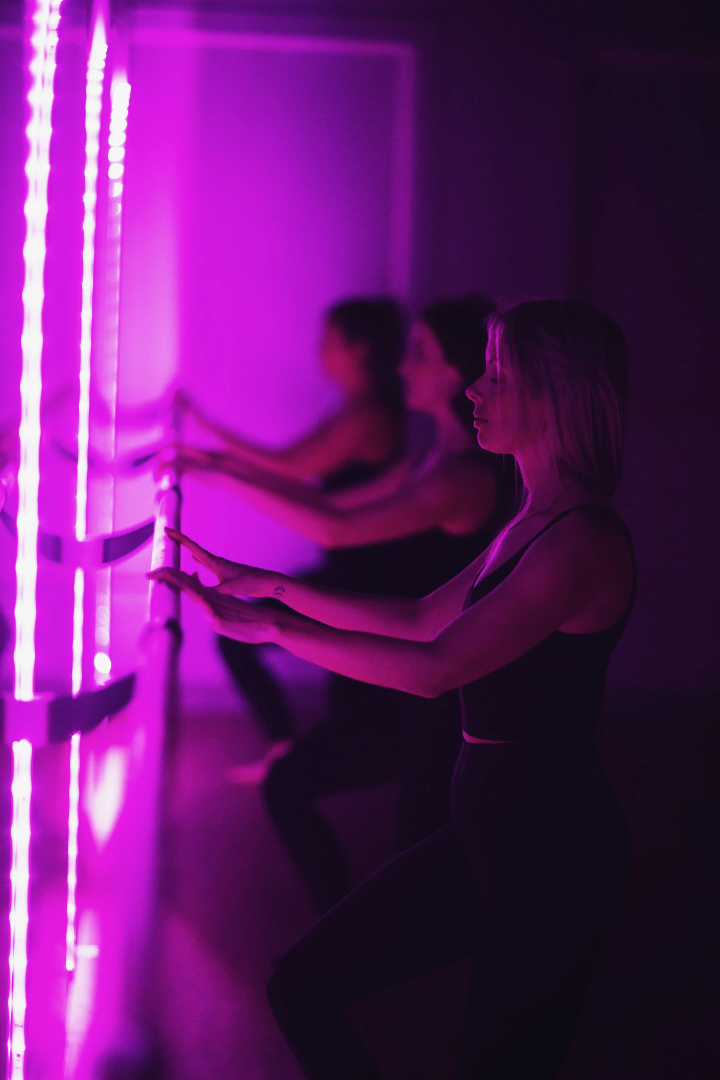 A fitness instructor leading a barre in a dark room illuminated with pink lighting, with participants in workout attire following her movements.