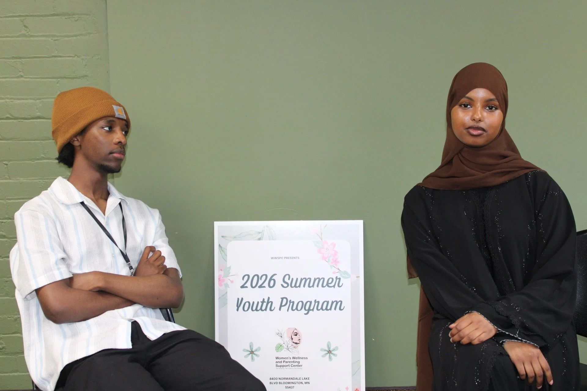 Two young adults standing on each side of a sign that reads '2026 Summer Youth Program,' with a green wall in the background.