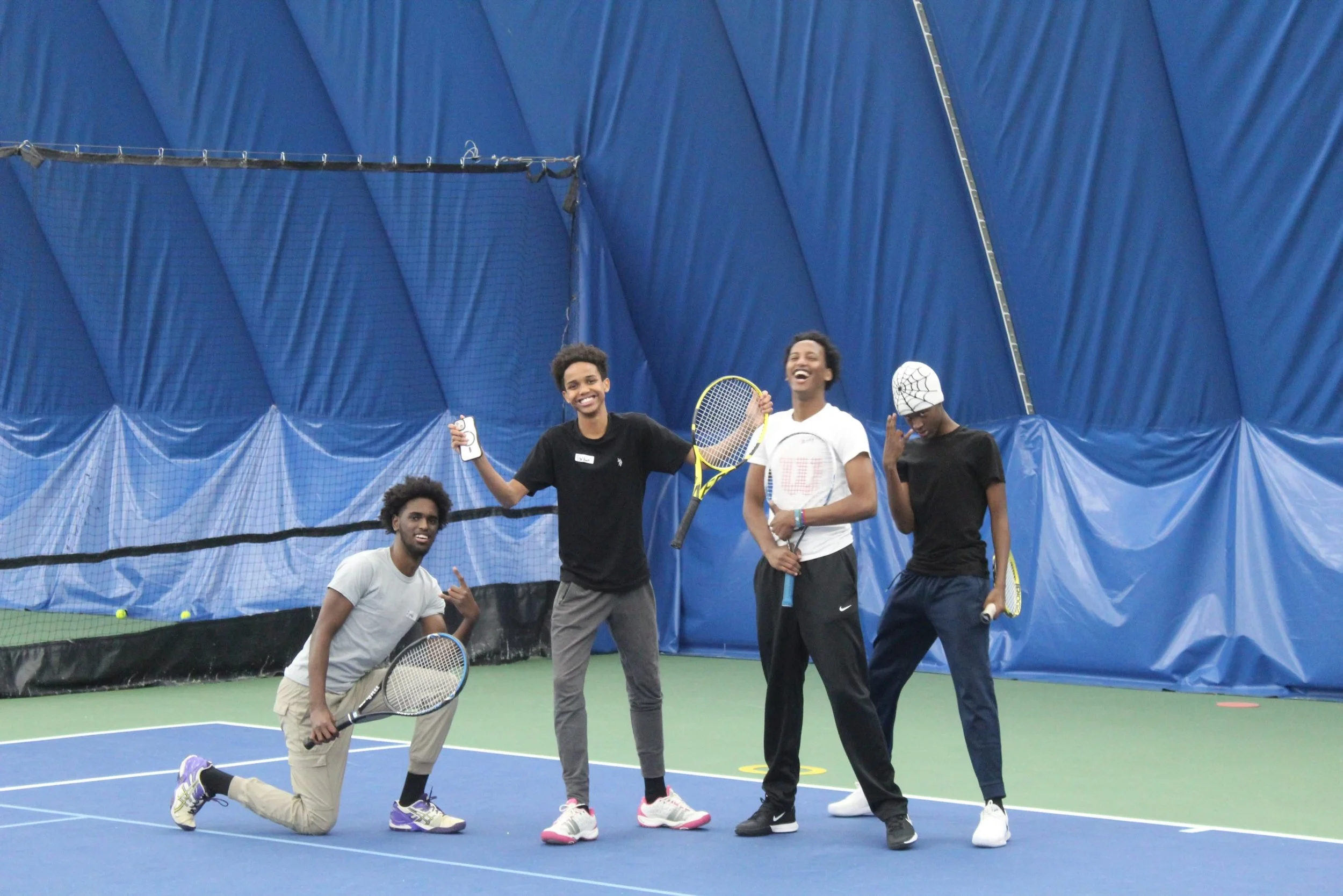 Group of five young men on an indoor tennis court posing playfully with tennis rackets and smiling.