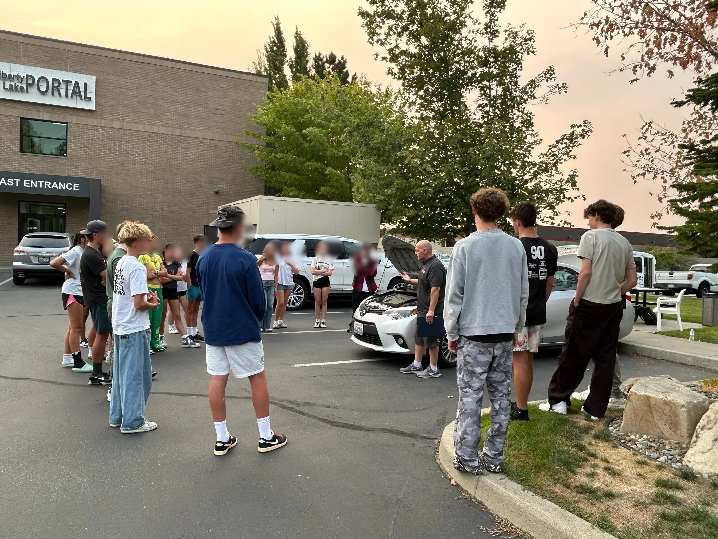 A group of teenagers and young adults are gathered outdoors in a parking lot, listening to a man with a clipboard by a silver car. The setting is near a building with a sign that reads 'Liberty Lake Portal' and 'East Entrance'.