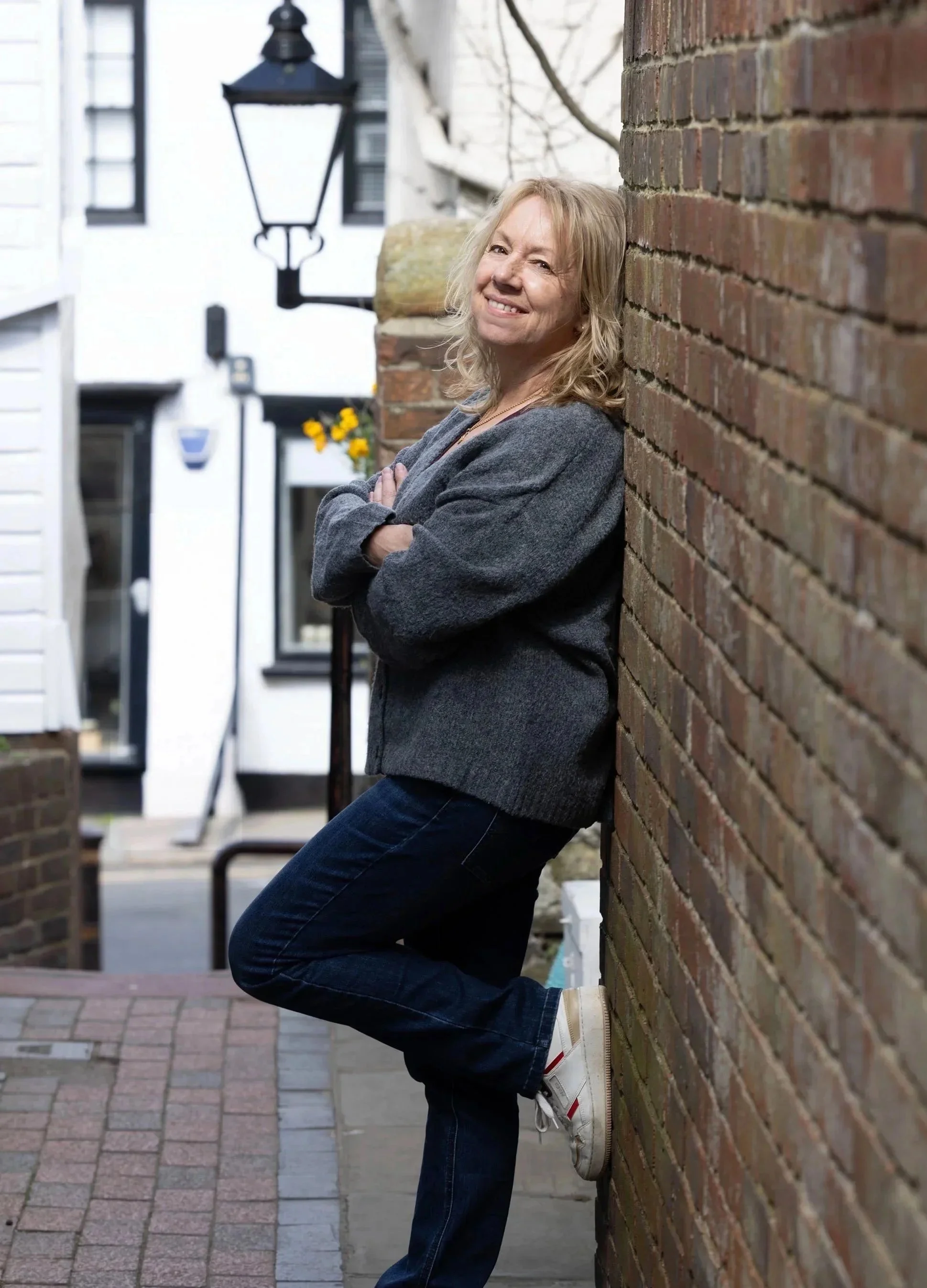 Faith Simpson, smiling and standing with her arms crossed, leaning against a brick wall outdoors in a residential area.