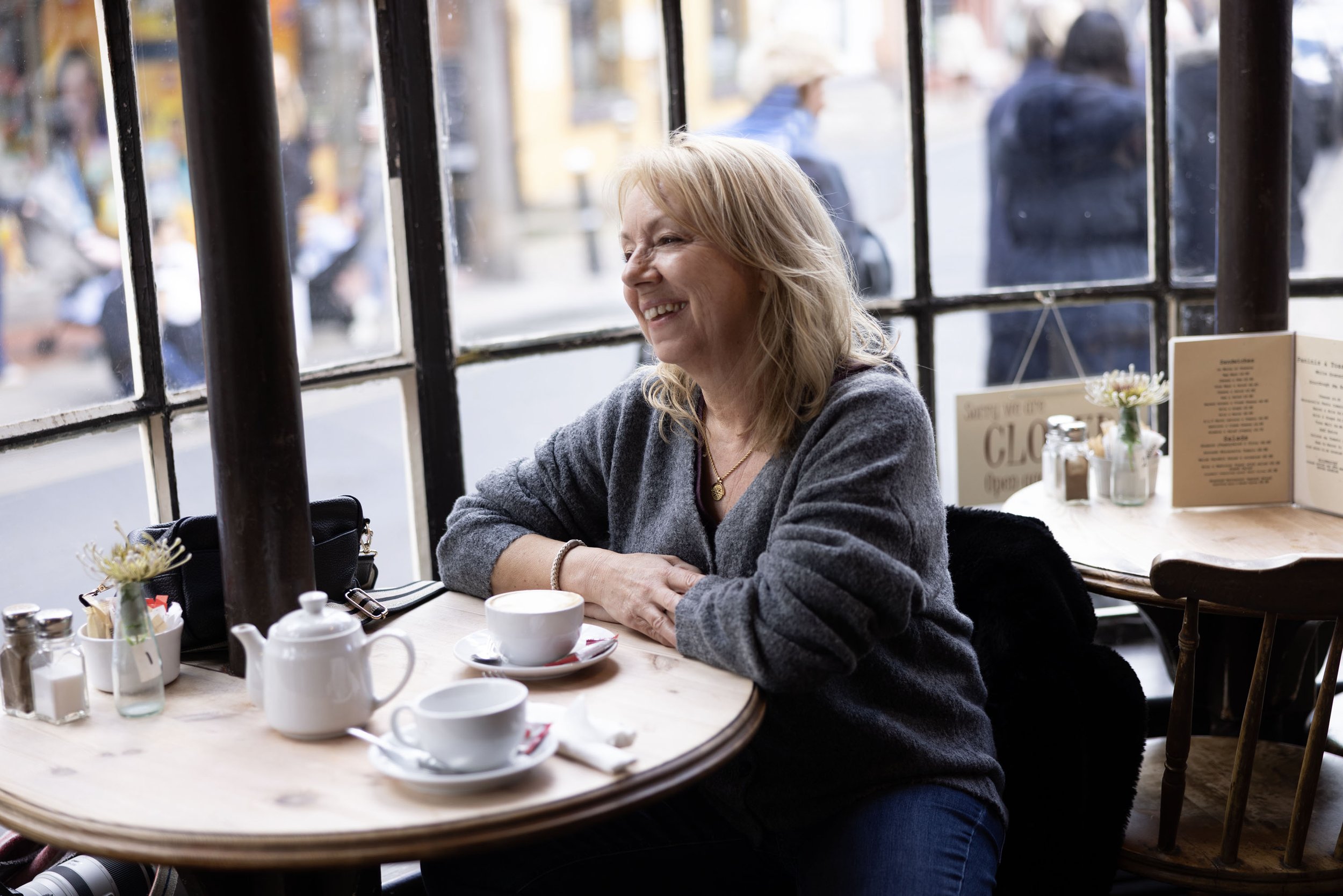 A woman with blonde hair smiling while sitting at a wooden table in a cafe by the window. She has a cup of coffee in front of her, and there are flowers and menu on the table. Outside, blurred pedestrians walk on the street.