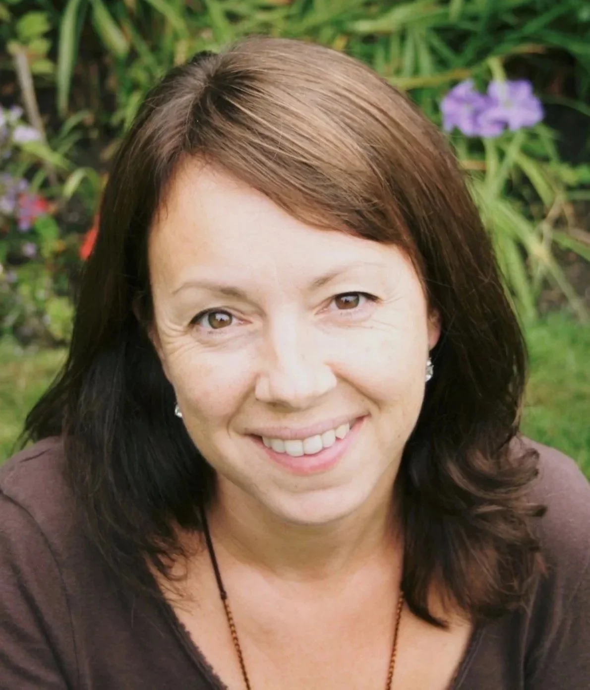 A woman with shoulder-length dark brown hair smiling outdoors with greenery and purple flowers in the background.
