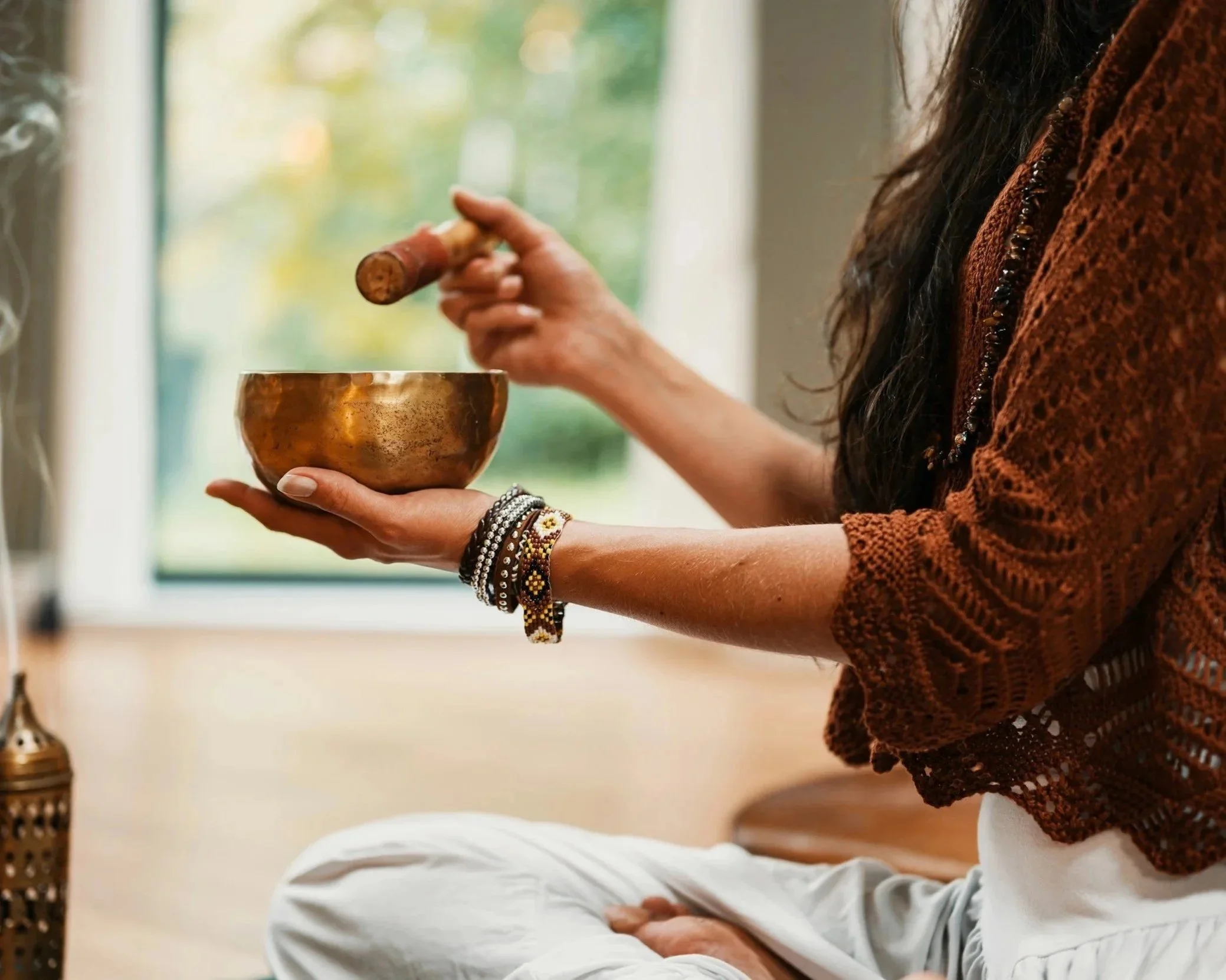 Person sitting cross-legged holding a metal singing bowl in one hand and a mallet in the other, preparing to play it, with a blurred window in the background.