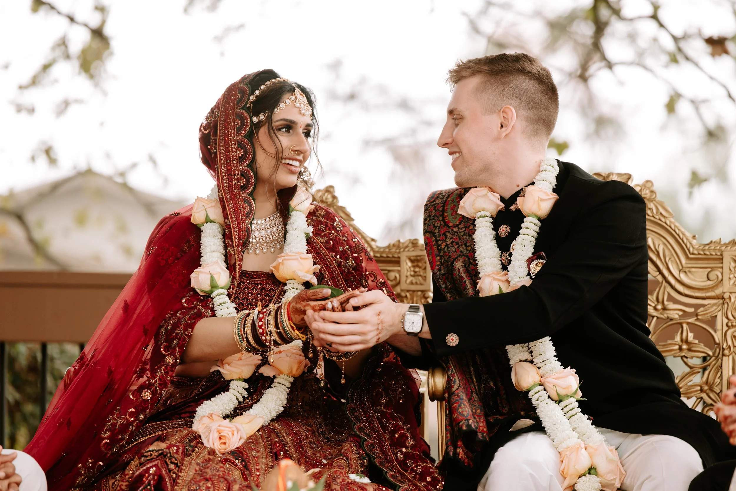 A bride and groom at a wedding ceremony, sitting on a decorated throne, exchanging flowers, wearing traditional outfits with floral garlands, and smiling at each other.